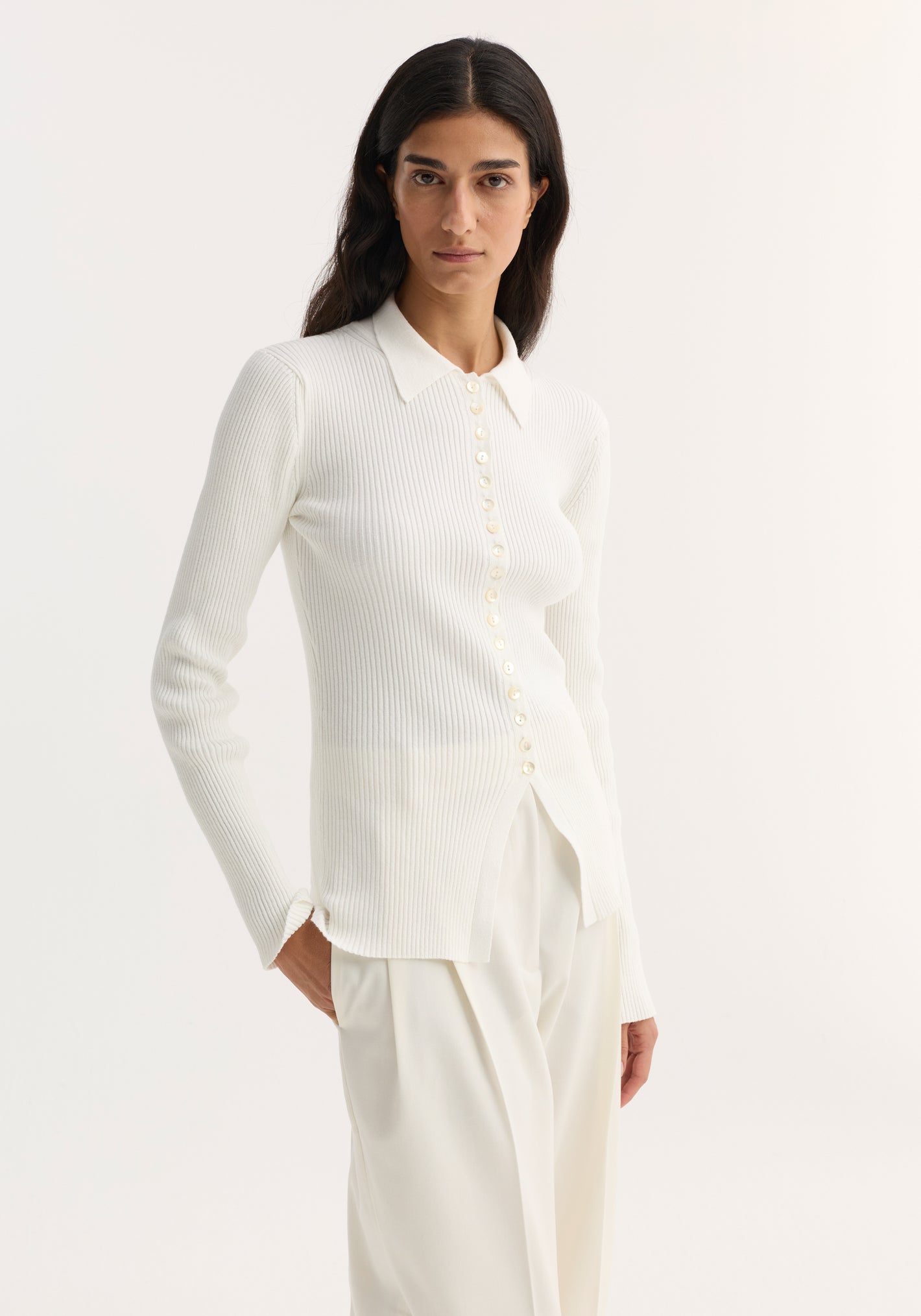 Elongated rib cardigan in off white with a pointed collar, button closure, and flared hem, paired with loose, high-waisted trousers.