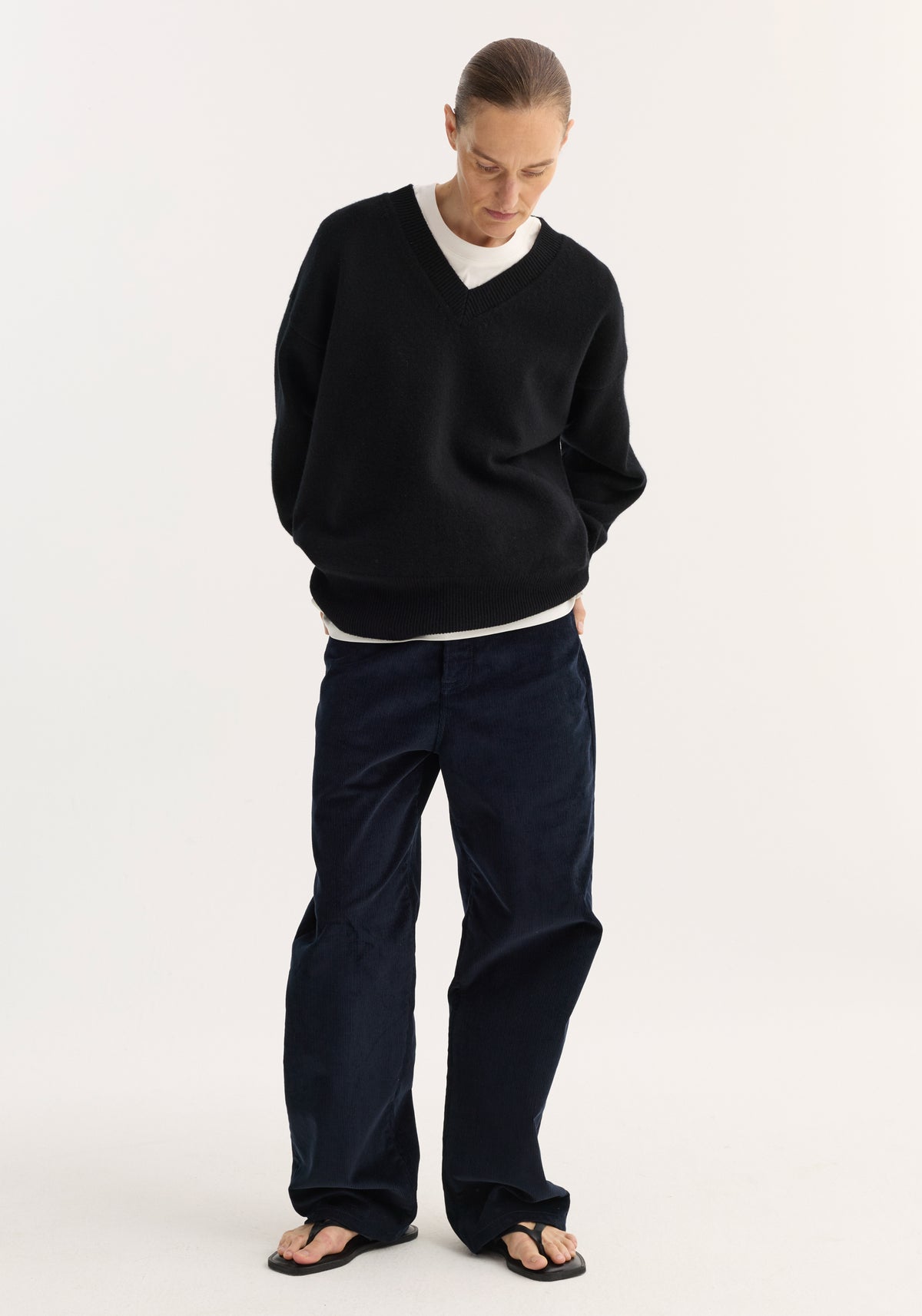 Model wearing a loose fitting V-neck knitted jumper | black over a white shirt, paired with dark wide-leg pants and black sandals, standing against a plain light background.