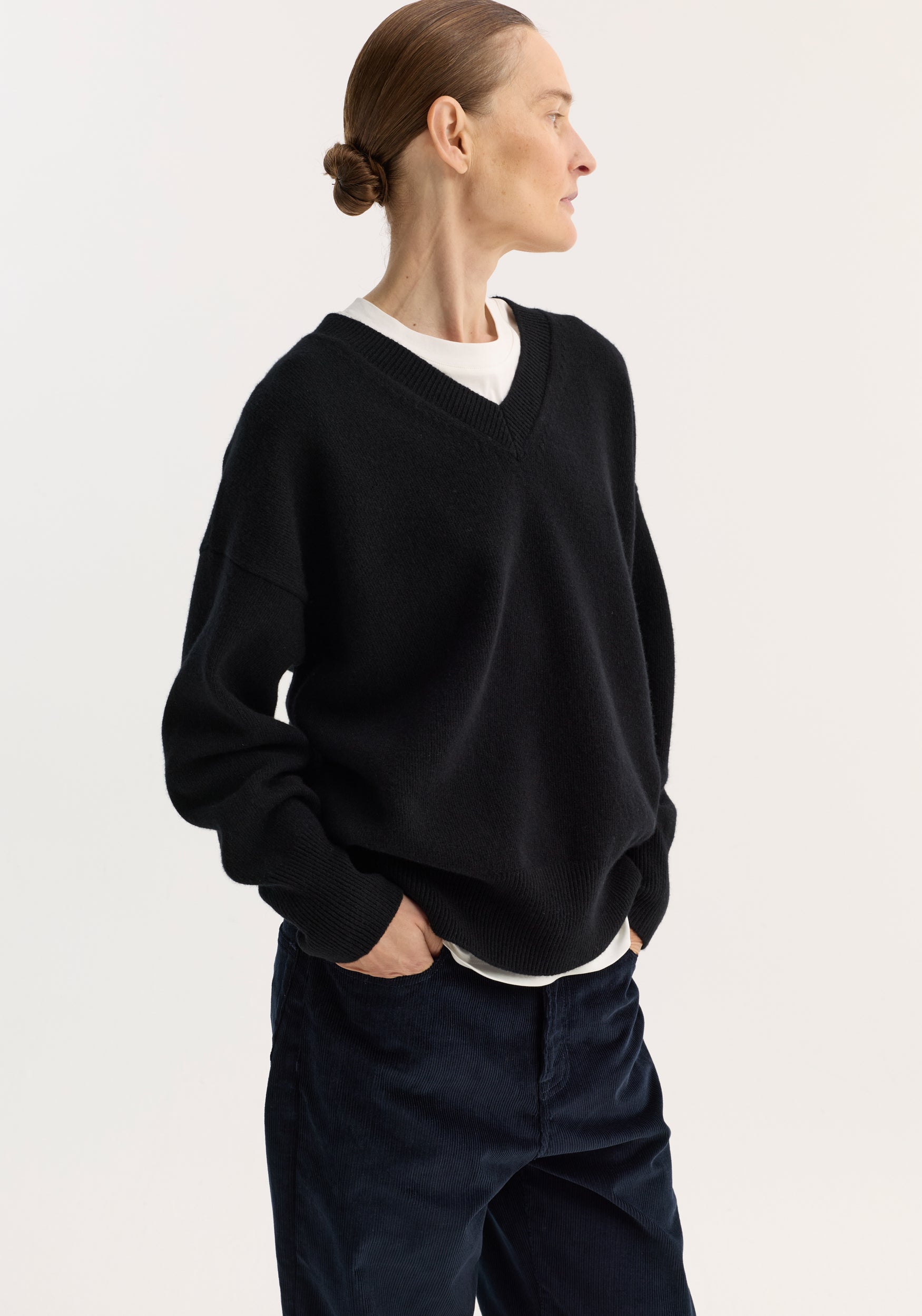 Woman wearing a black V-neck knitted jumper | black over a white shirt, paired with dark pants, standing sideways with hands in pockets against a plain background.