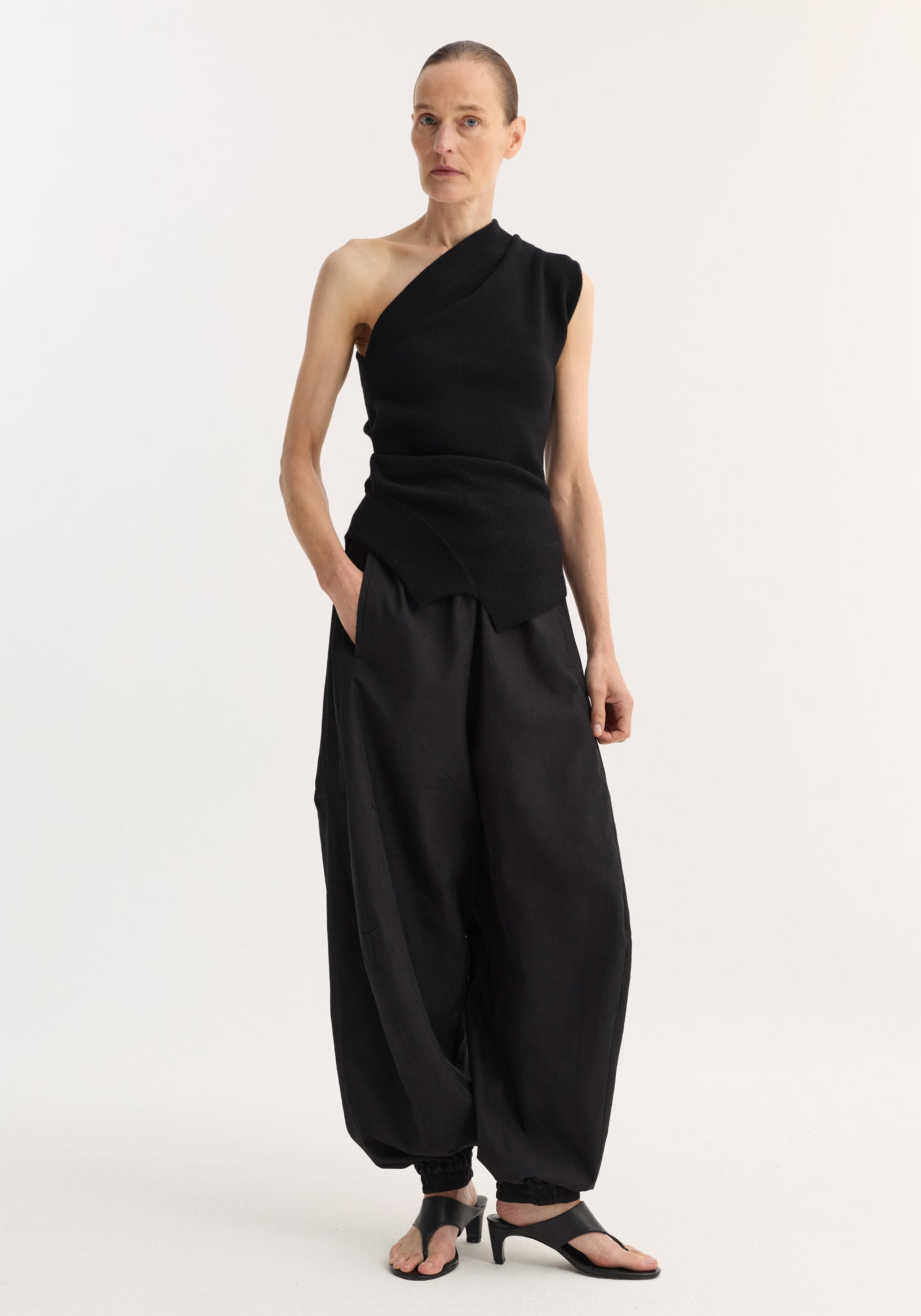 A model wearing a sleek off-shoulder top | black paired with loose-fitting black pants and black sandals, standing against a plain neutral background.