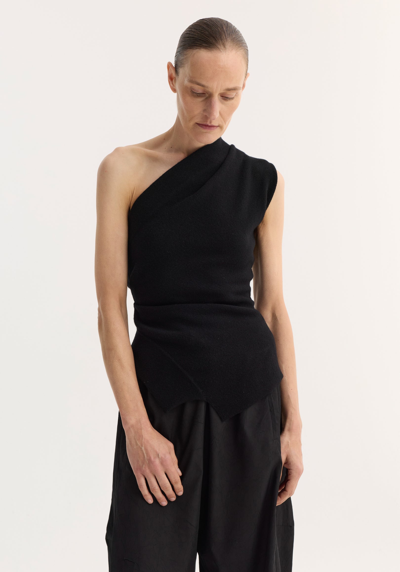 Woman wearing a black off-shoulder top | black with one sleeve and an asymmetrical hem, paired with loose black pants against a plain light background.