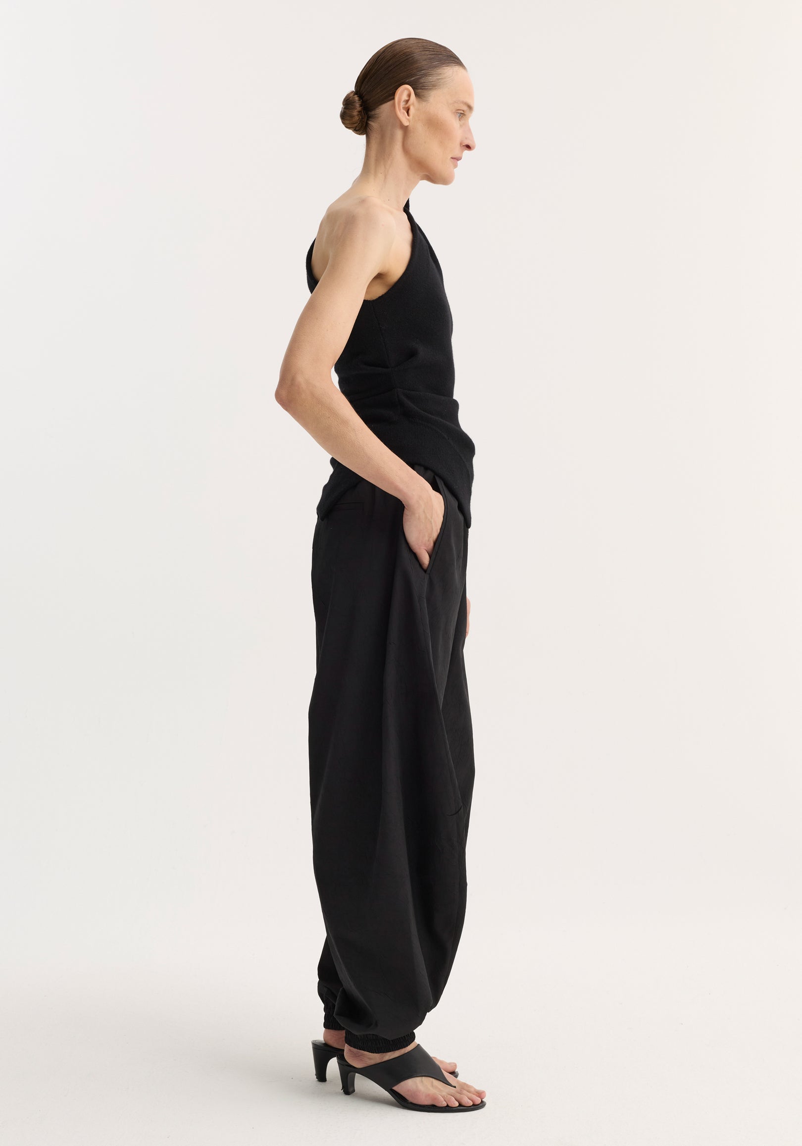 Side view of a woman wearing an off-shoulder top | black paired with loose black pants and black heeled sandals, standing against a plain light background.