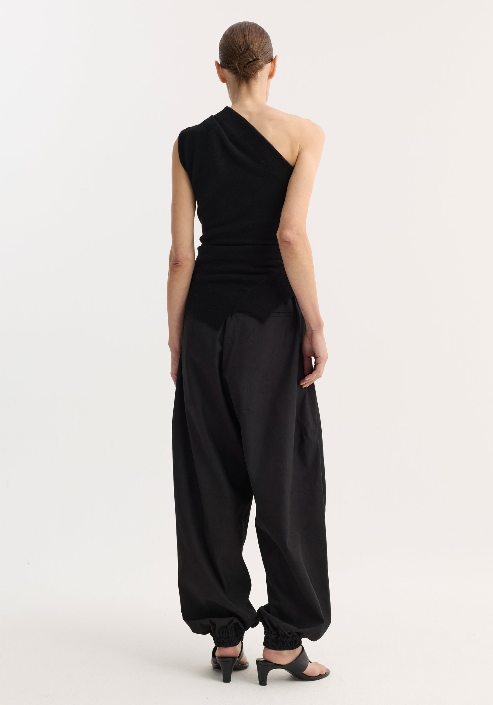 Woman seen from the back wearing an off-shoulder top | black paired with loose black pants and black open-toe heels, standing against a plain white background.