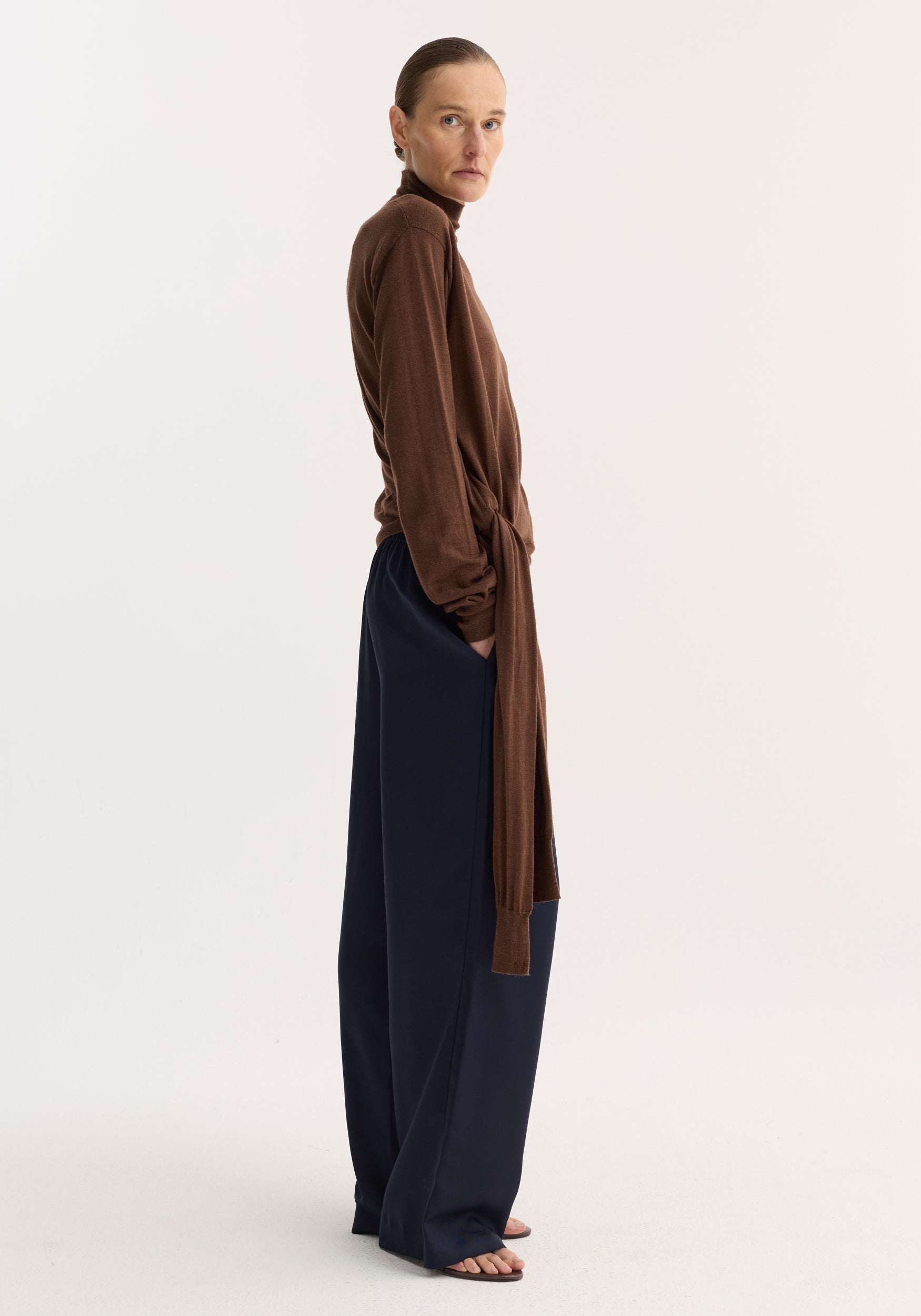 Model wearing wide-leg silk trousers with elasticated waistband and drawstring, showcasing relaxed drape and fluidity, styled with a brown turtleneck sweater.