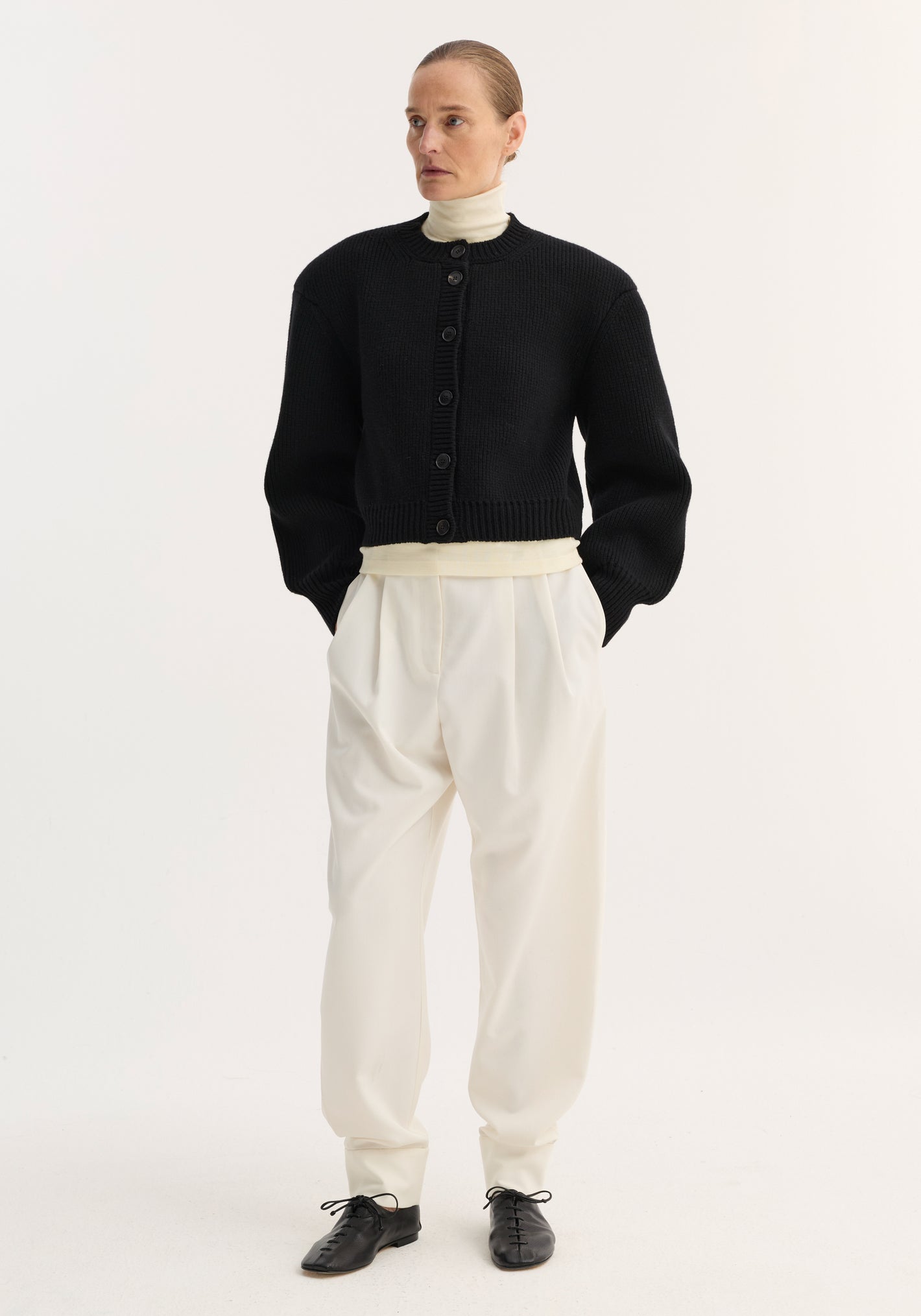 Woman wearing Barrel leg trousers in off-white, high-waisted with double pleats and ankle cuffs, styled with a black cardigan and cream turtleneck.