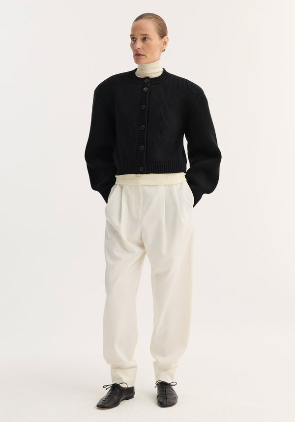 Woman wearing Barrel leg trousers in off-white, high-waisted with double pleats and ankle cuffs, styled with a black cardigan and cream turtleneck.