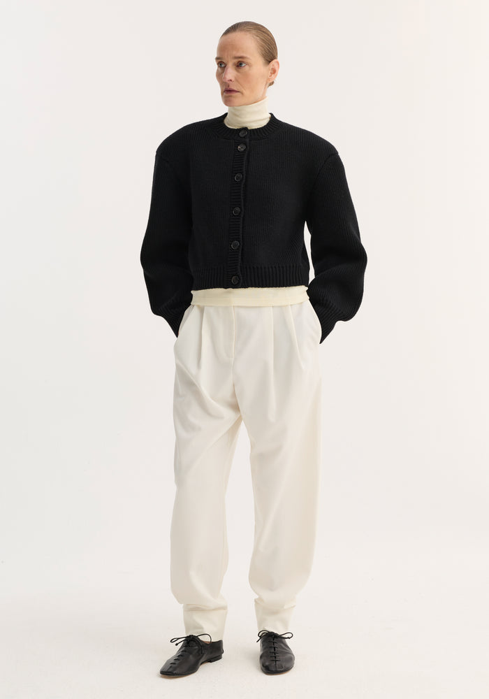 Woman wearing Barrel leg trousers in off-white, high-waisted with double pleats and ankle cuffs, styled with a black cardigan and cream turtleneck.