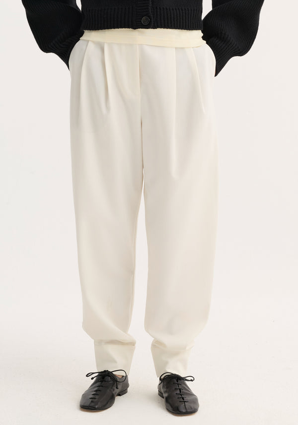 Barrel leg trousers in off-white wool blend, high-waisted with single pleat, tapering to ankle cuffs, paired with black flats against a light background.