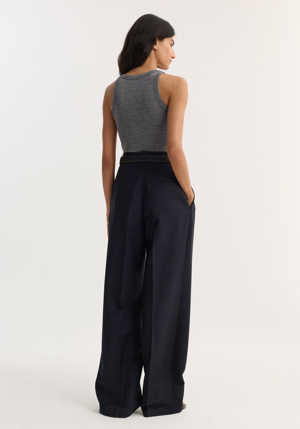 Back view of model wearing mid-waist tailored trousers with wide legs, belted waist, and double pleats, hands in pockets for a relaxed look.