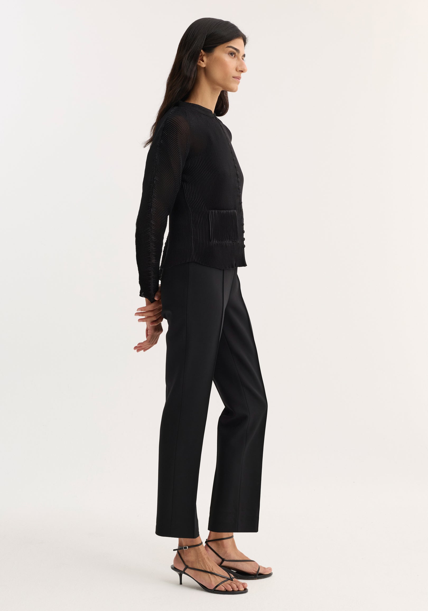 Woman wearing a black plissé cardigan | black featuring long sleeves and a textured fabric, paired with black tailored pants and black strappy heeled sandals, standing in profile.