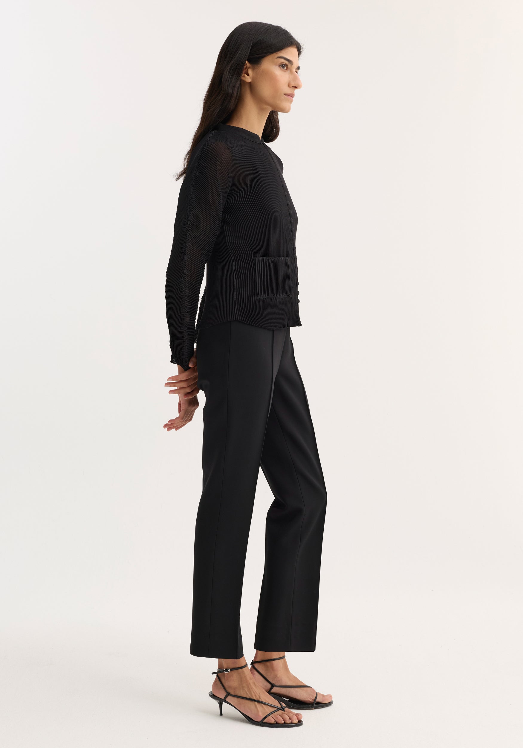 Woman wearing a black plissé cardigan | black featuring long sleeves and a textured fabric, paired with black tailored pants and black strappy heeled sandals, standing in profile.