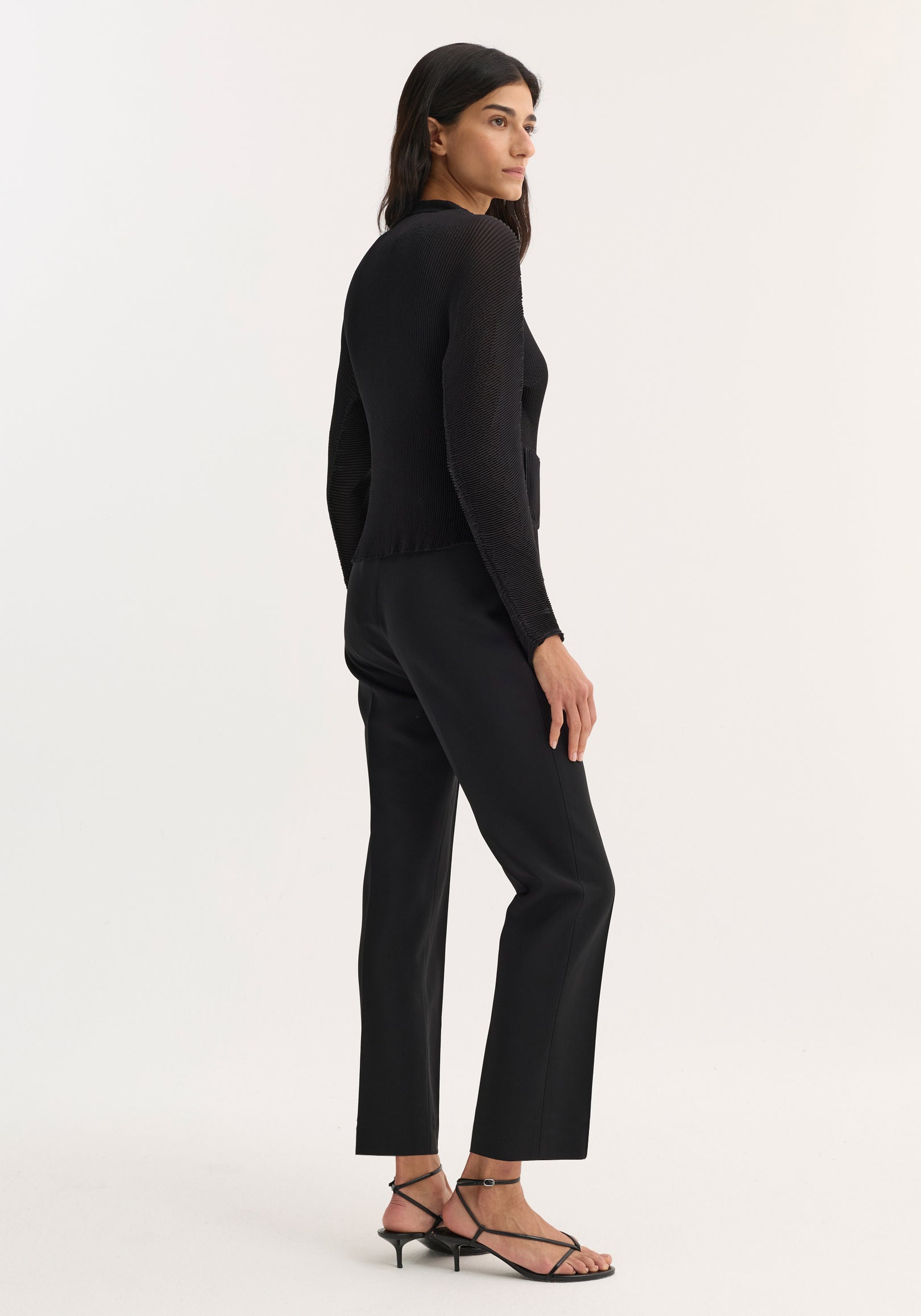 Woman wearing a fitted Plissé cardigan | black with long sleeves, paired with black straight-leg pants and strappy black sandals, standing against a light background.