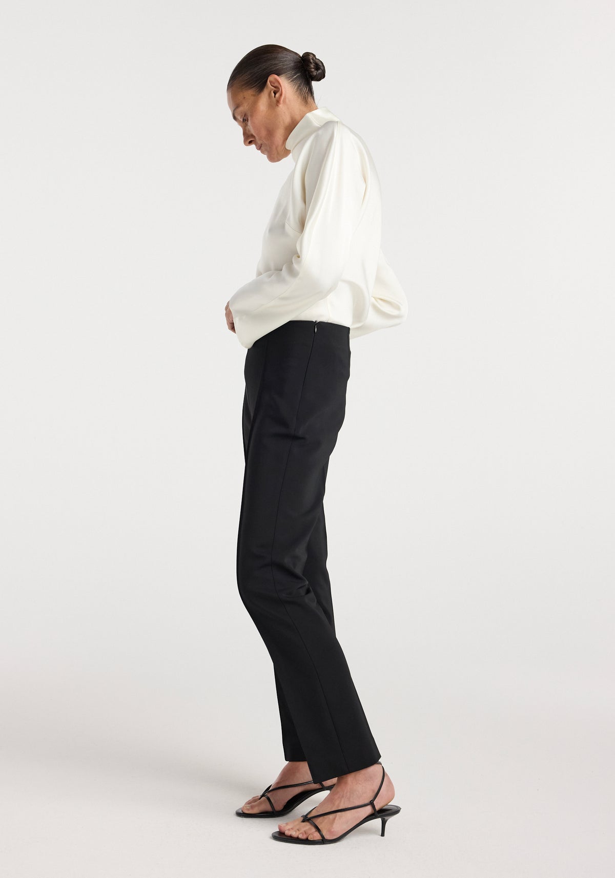 Woman wearing black Cigarette leg trousers | black paired with a white long-sleeve top and black strappy low-heeled sandals, standing in profile against a plain background.