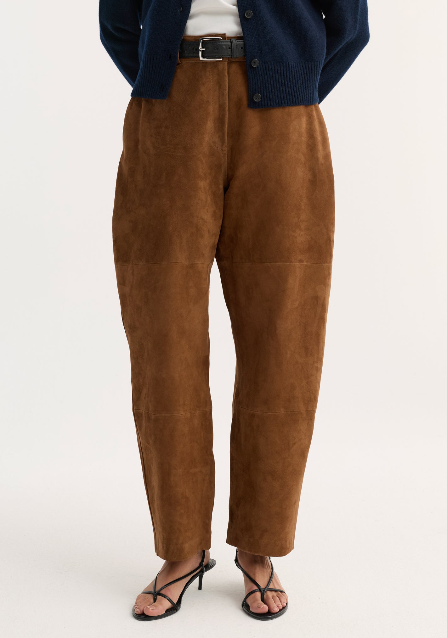 Model wearing Sculptural suede trousers in tobacco, showcasing high-waisted, barrel-leg design with panel seam detailing. Paired with a black leather belt and strappy heeled sandals.
