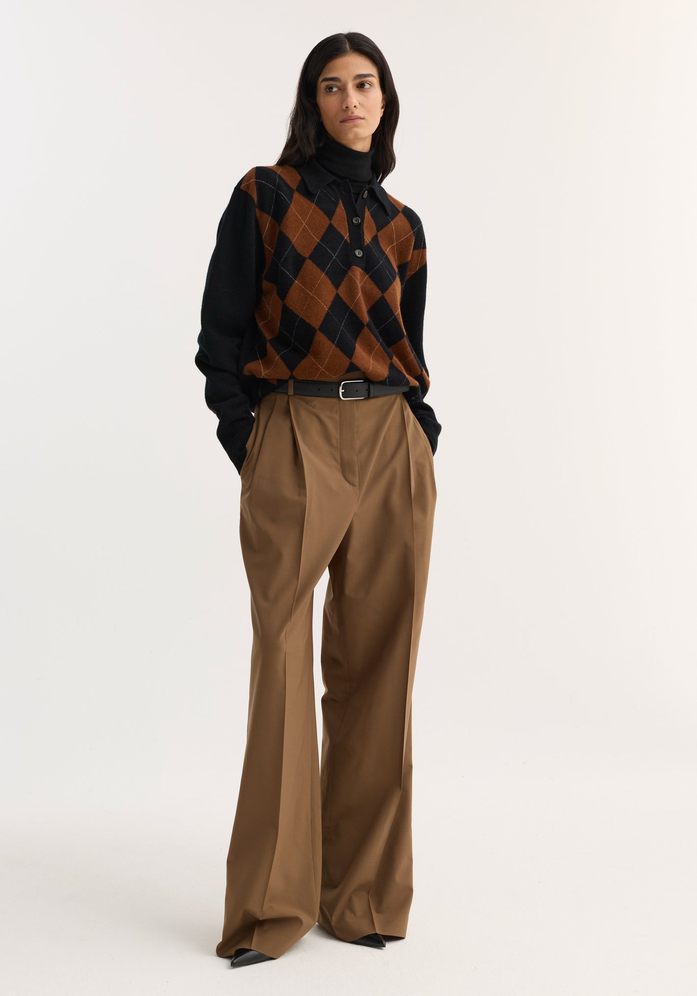 Model wearing Mid-waist tailored trousers in mocha with a wide leg, double pleats, and a structured waistband, styled with a black turtleneck and argyle sweater.