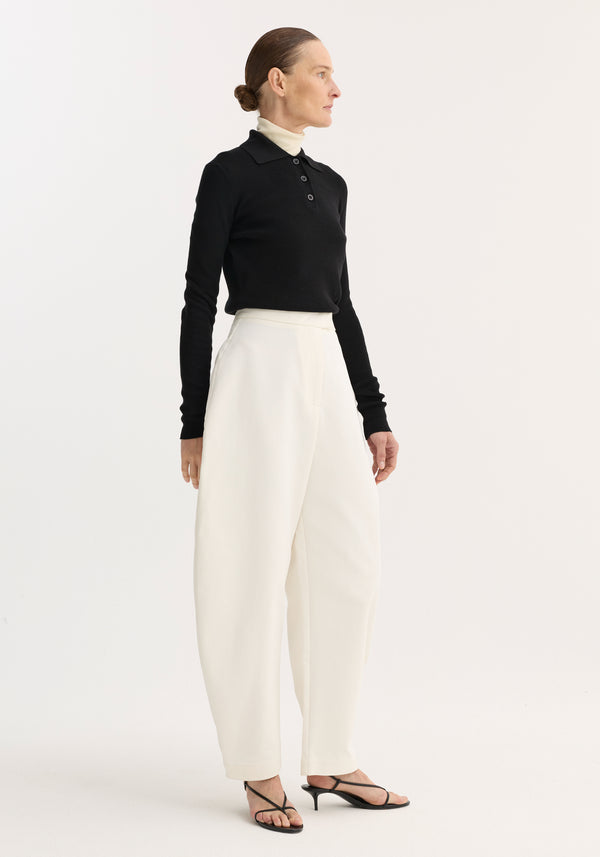 Woman models Sculptural trousers in paper white, featuring a high waist and bold balloon leg, styled with black top and heeled sandals.
