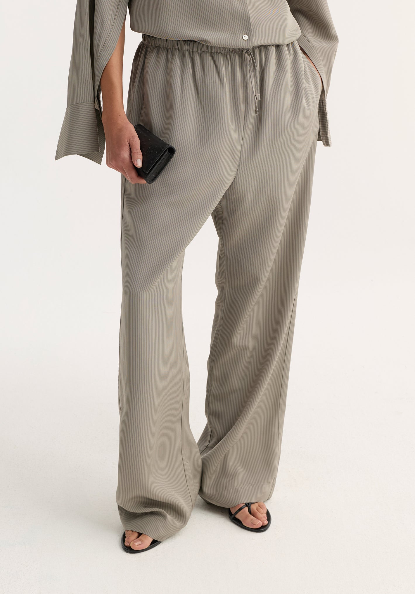 Relaxed fit trousers in light grey stripe, featuring a wide leg, drawstring waistband, and side seam pockets, styled with black sandals and a leather clutch.