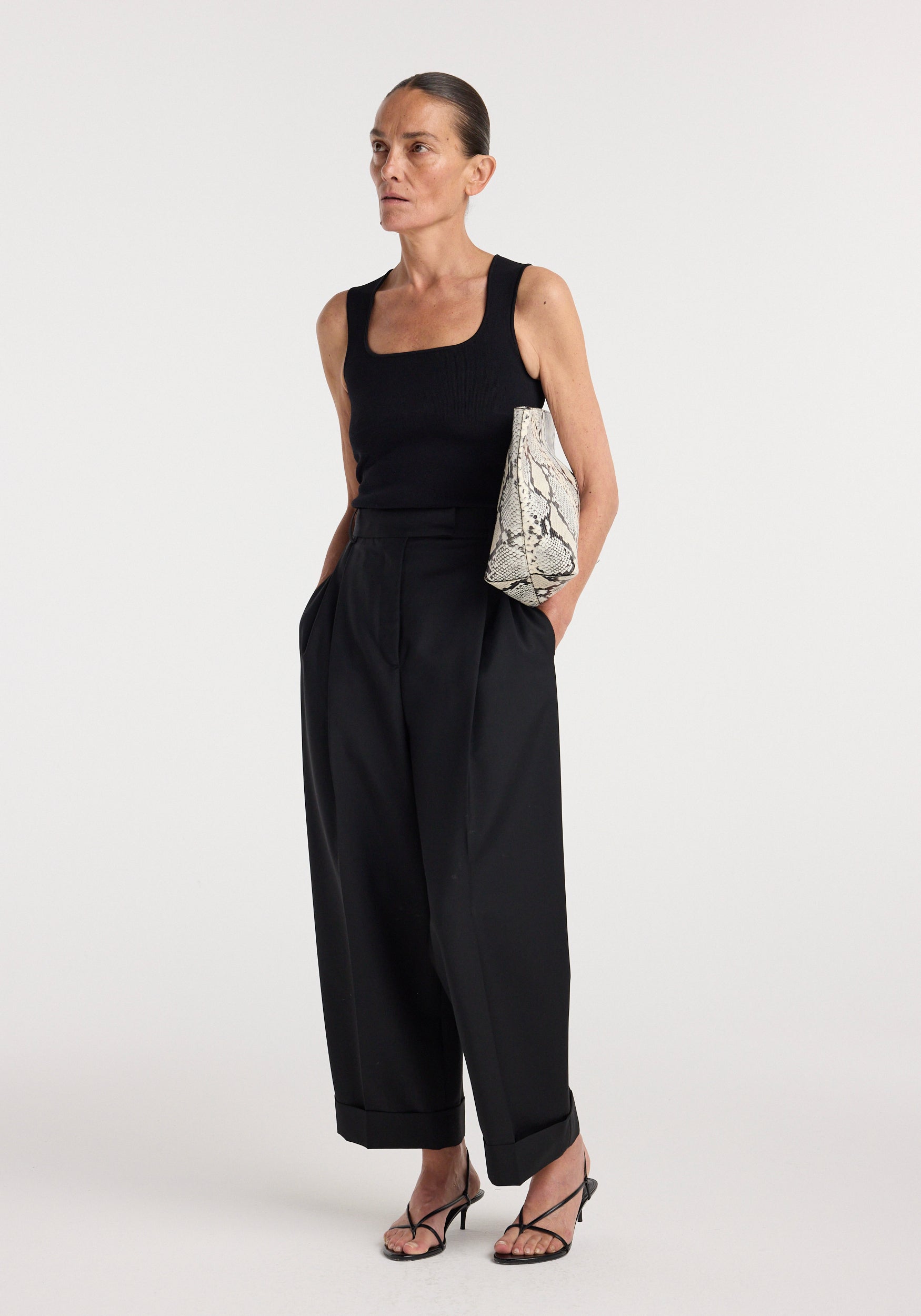 Woman wearing a Bustier shaped knitted top | black paired with high-waisted wide-leg black trousers and black strappy heels, holding a snakeskin-patterned clutch bag.
