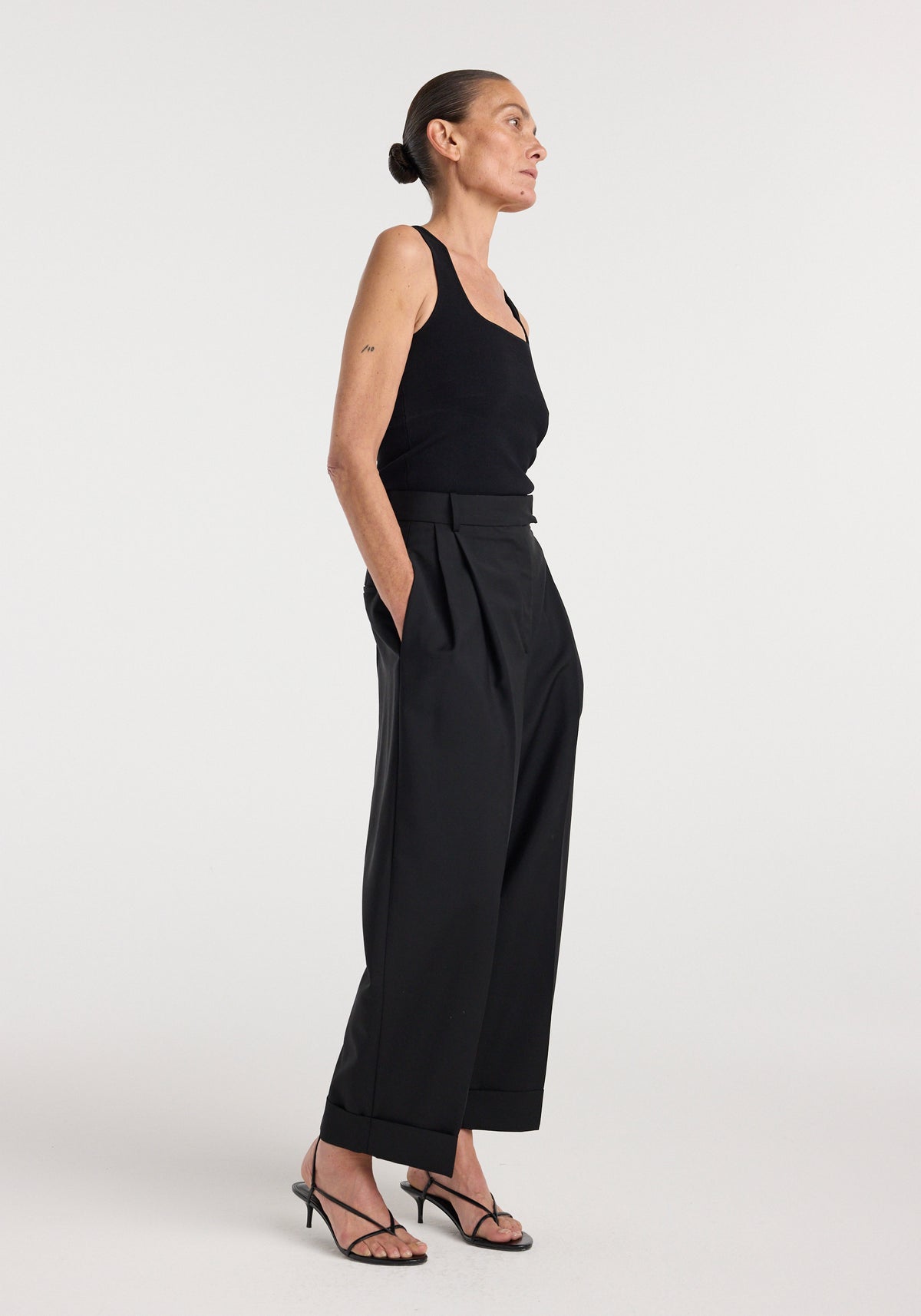 Woman wearing a black bustier shaped knitted top | black with high-waisted, wide-leg black trousers and black strappy heels against a plain background.
