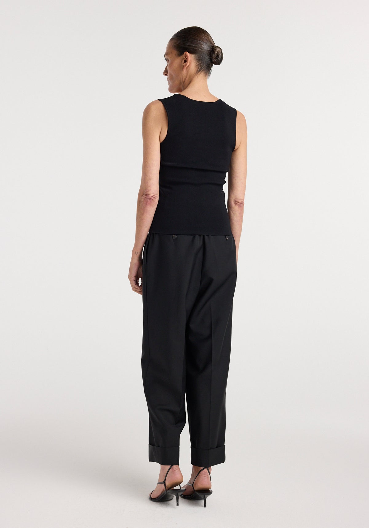 Woman seen from the back wearing a sleeveless Bustier shaped knitted top | black paired with loose black trousers and black heeled sandals against a plain background.