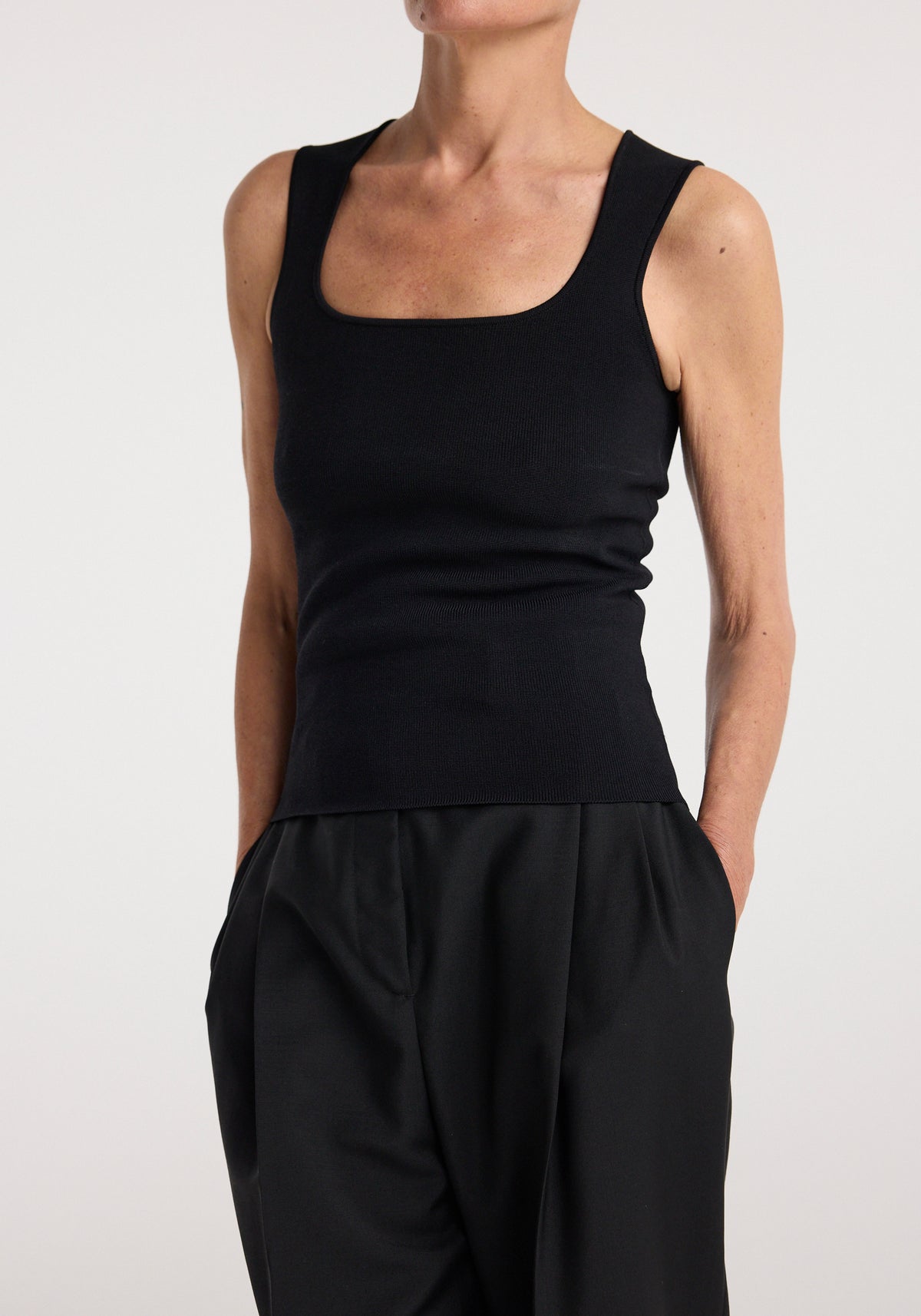 Woman wearing a fitted black sleeveless top with a square neckline and a bustier shaped knitted top | black paired with loose black pants against a plain background.