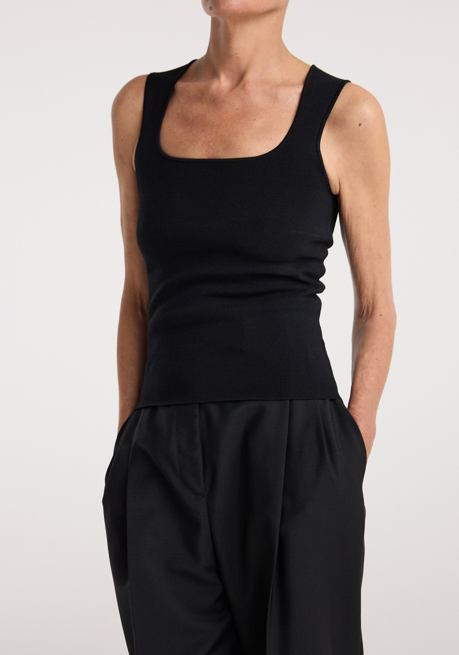 Woman wearing a fitted black sleeveless top with a square neckline and a bustier shaped knitted top | black paired with loose black pants against a plain background.