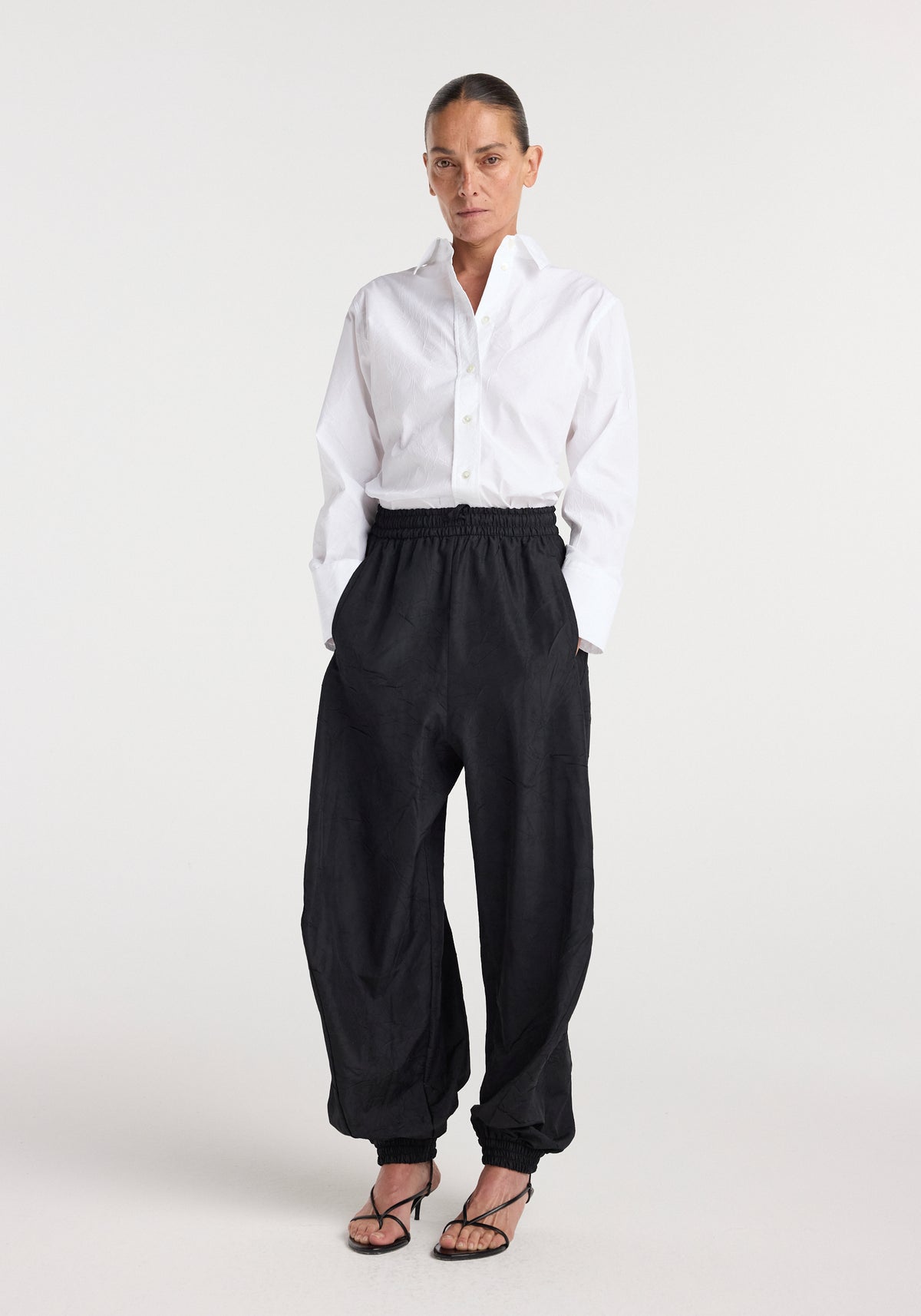 Woman wearing black Balloon track pants | black with elastic waistband and cuffs, paired with a white button-up shirt and black strappy heels, standing against a plain background.