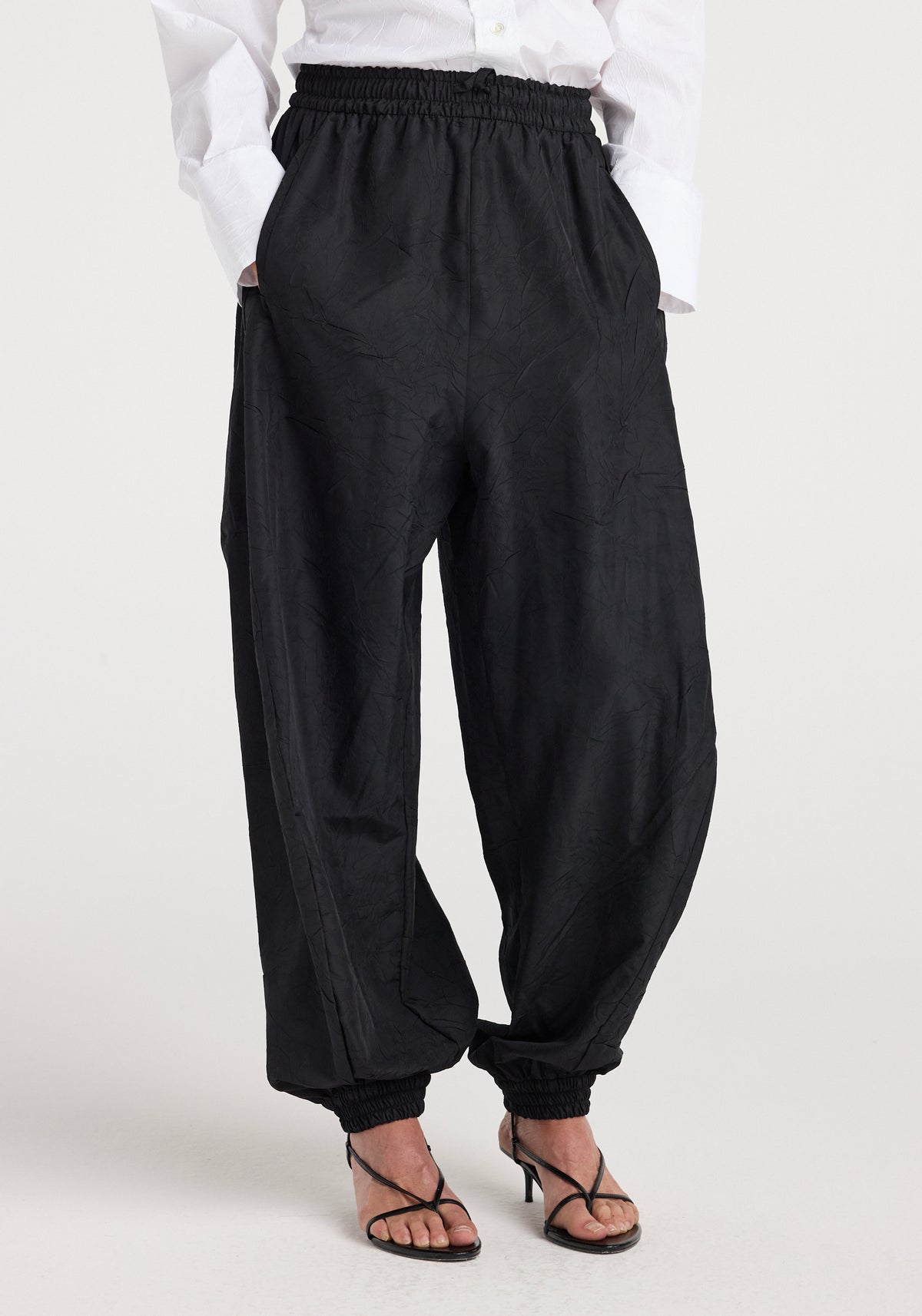 A person wearing black Balloon track pants | black with an elastic waistband and ankle cuffs, paired with black strappy heeled sandals and a white long-sleeve shirt.