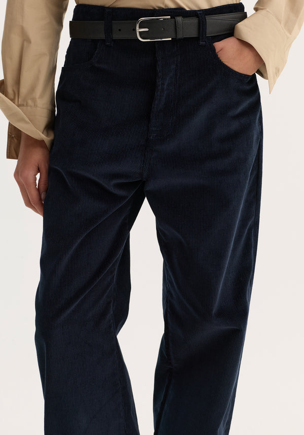 Close-up of Relaxed corduroy trousers | night, featuring navy corduroy, loose leg, denim-style details; worn with a black belt and beige shirt.