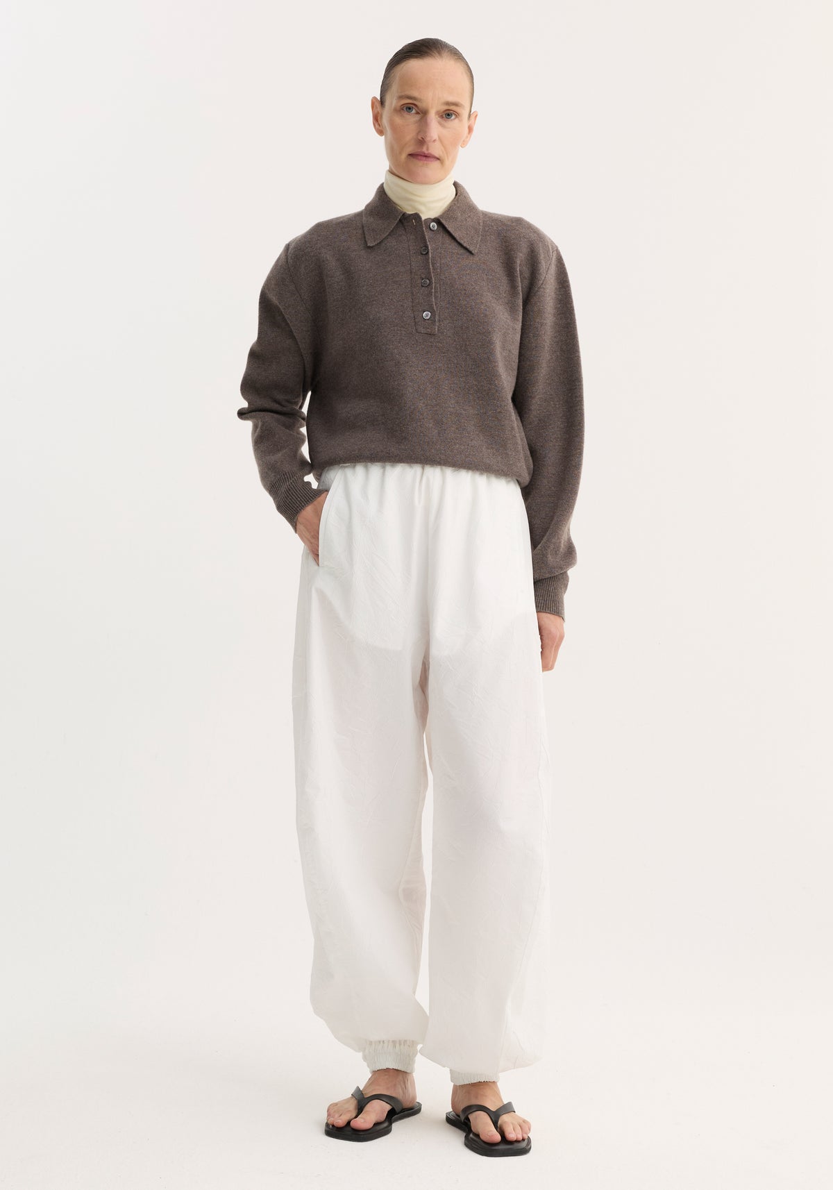 Model wearing Balloon track pants in optic white with a crinkled texture, elasticated waistband, and voluminous legs, paired with a brown polo sweater.
