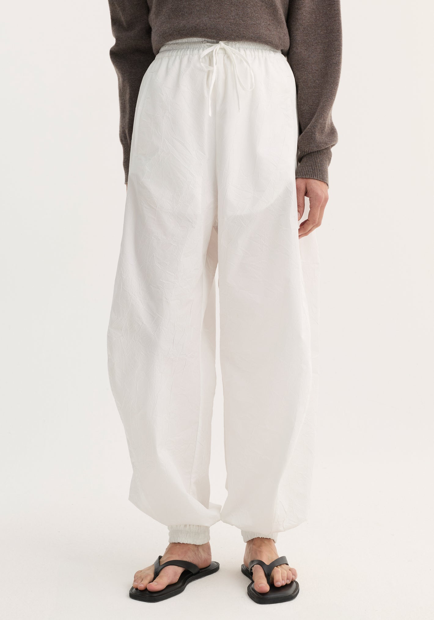 Balloon track pants in optic white with elasticated waistband and cuffs, featuring a crinkled texture and front welt pockets, paired with black sandals.