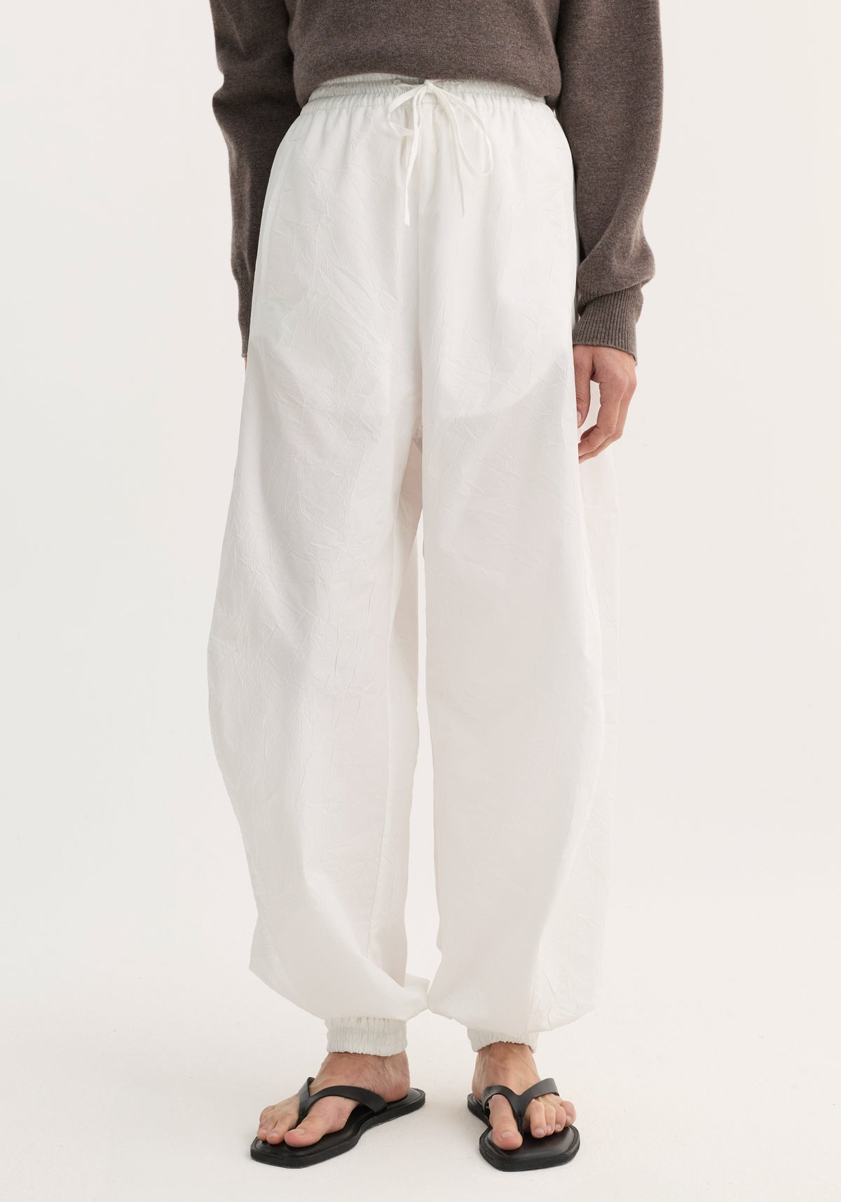 Balloon track pants in optic white with elasticated waistband and cuffs, featuring a crinkled texture and front welt pockets, paired with black sandals.