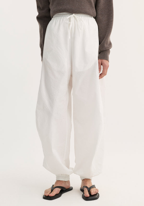 Balloon track pants in optic white with elasticated waistband and cuffs, featuring a crinkled texture and front welt pockets, paired with black sandals.