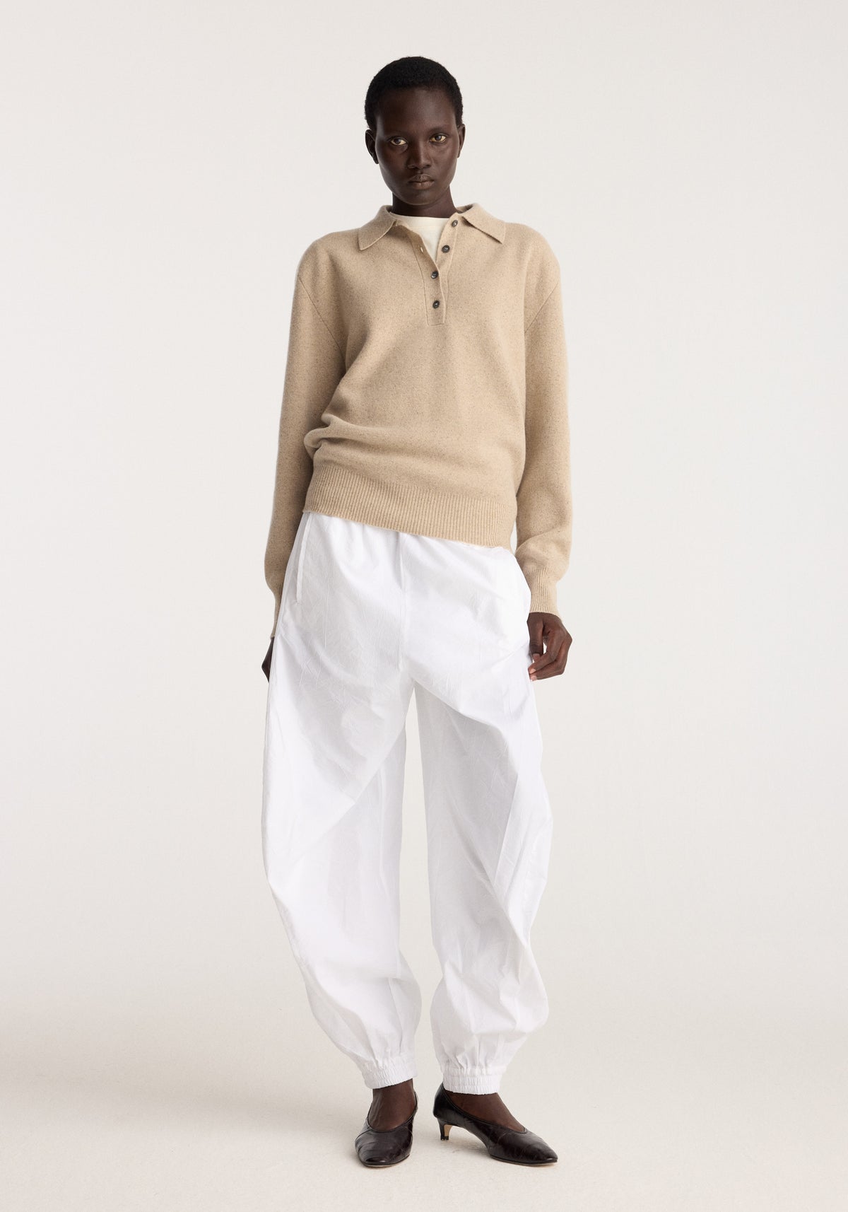 Model wearing Balloon track pants | optic white, relaxed white pants with elastic cuffs and a balloon silhouette styled with a beige knit polo sweater.