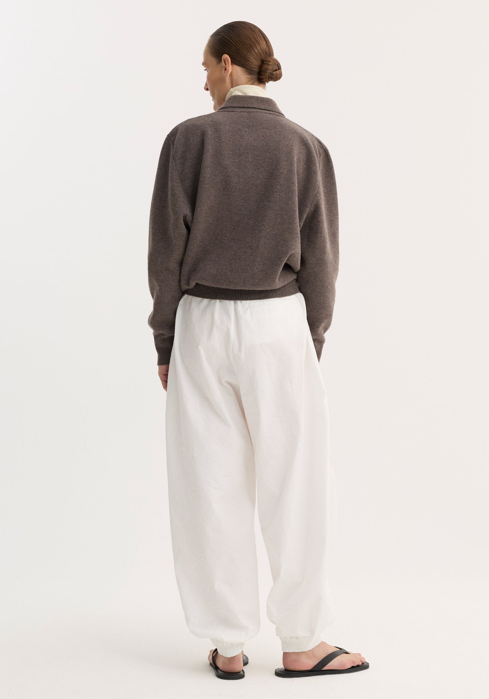 Back view of model in Balloon track pants | optic white, featuring crinkled texture, elastic waistband, voluminous legs, and welt pockets, paired with a muted sweater.
