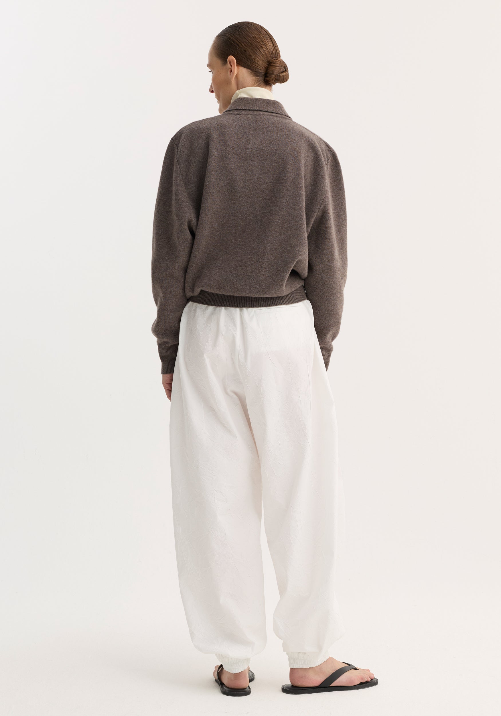 Back view of model in Balloon track pants | optic white, featuring crinkled texture, elastic waistband, voluminous legs, and welt pockets, paired with a muted sweater.
