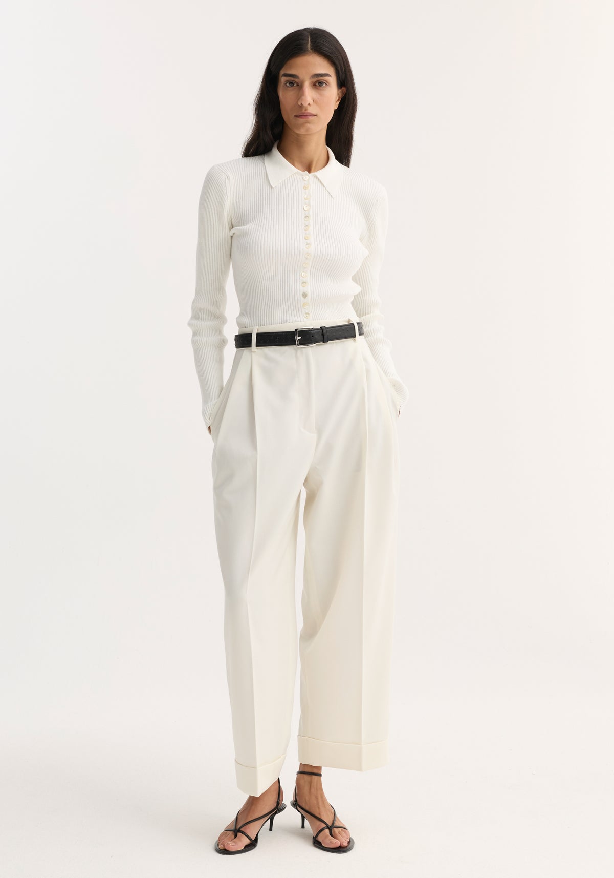 Woman wearing high-waist turn-up trousers with sharp pleats, paired with a ribbed knit top, black belt, and strappy sandals.
