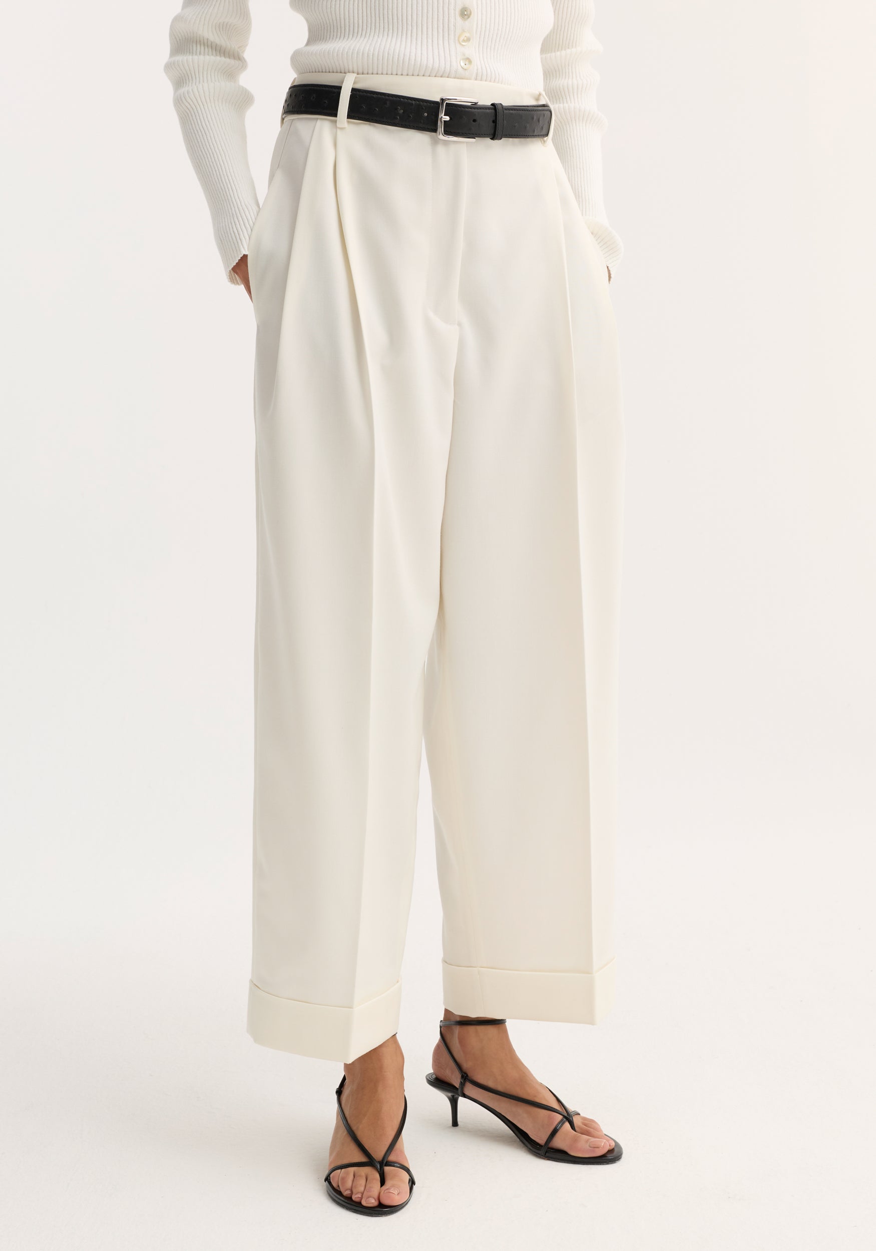 High-waist turn-up trousers in off-white, featuring wide legs, subtle pleats, a deep cuff, and side pockets, styled with a black belt and sandals.