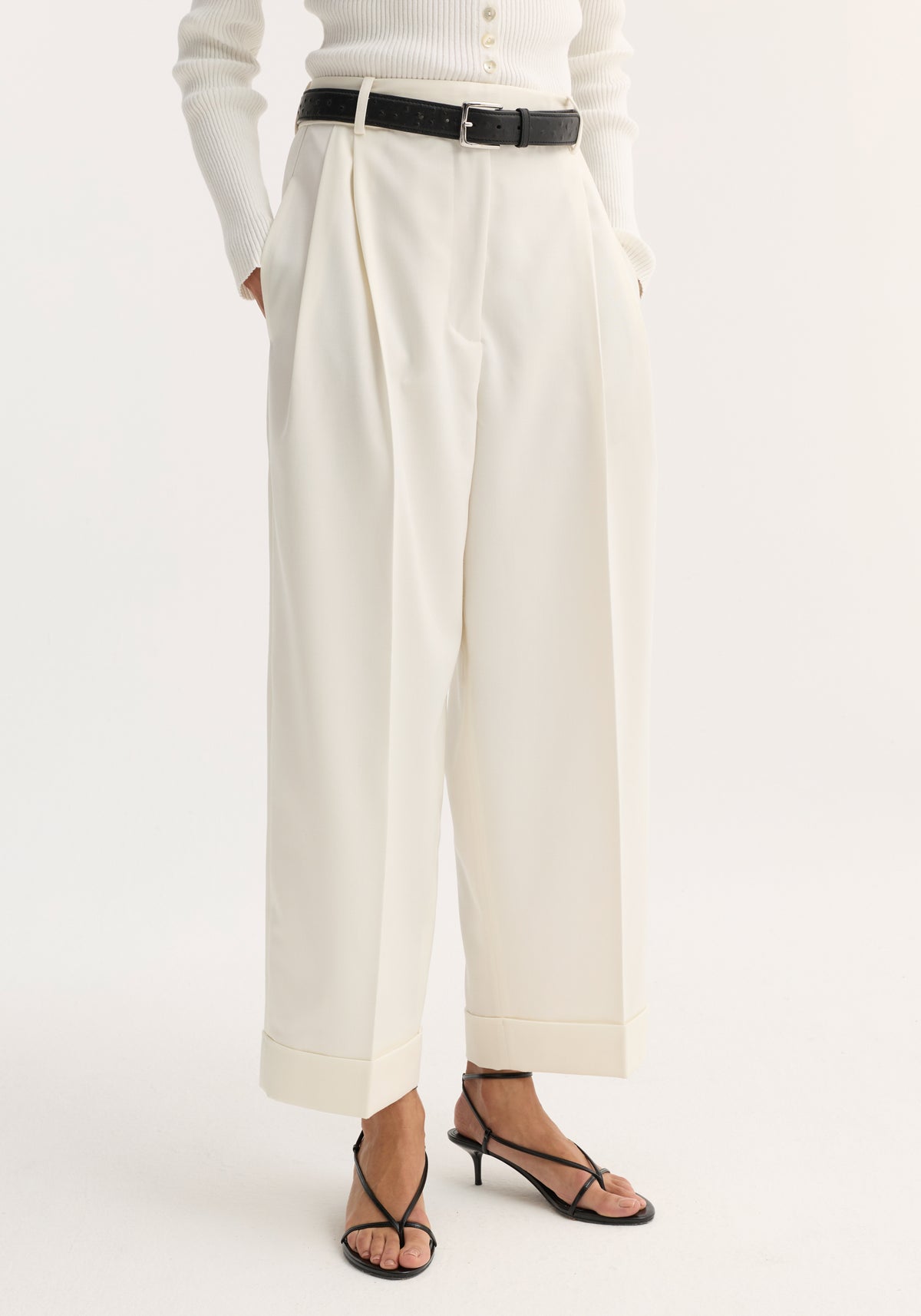 High-waist turn-up trousers in off-white, featuring wide legs, subtle pleats, a deep cuff, and side pockets, styled with a black belt and sandals.