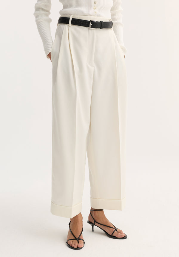 High-waist turn-up trousers in off-white, featuring wide legs, subtle pleats, a deep cuff, and side pockets, styled with a black belt and sandals.