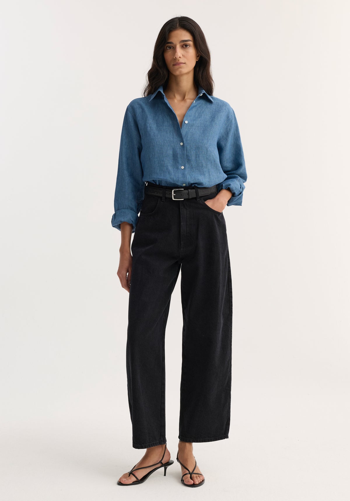 Woman wearing a blue button-up shirt paired with black Berber - Barrel leg denim | black jeans, styled with a black belt and black strappy sandals.