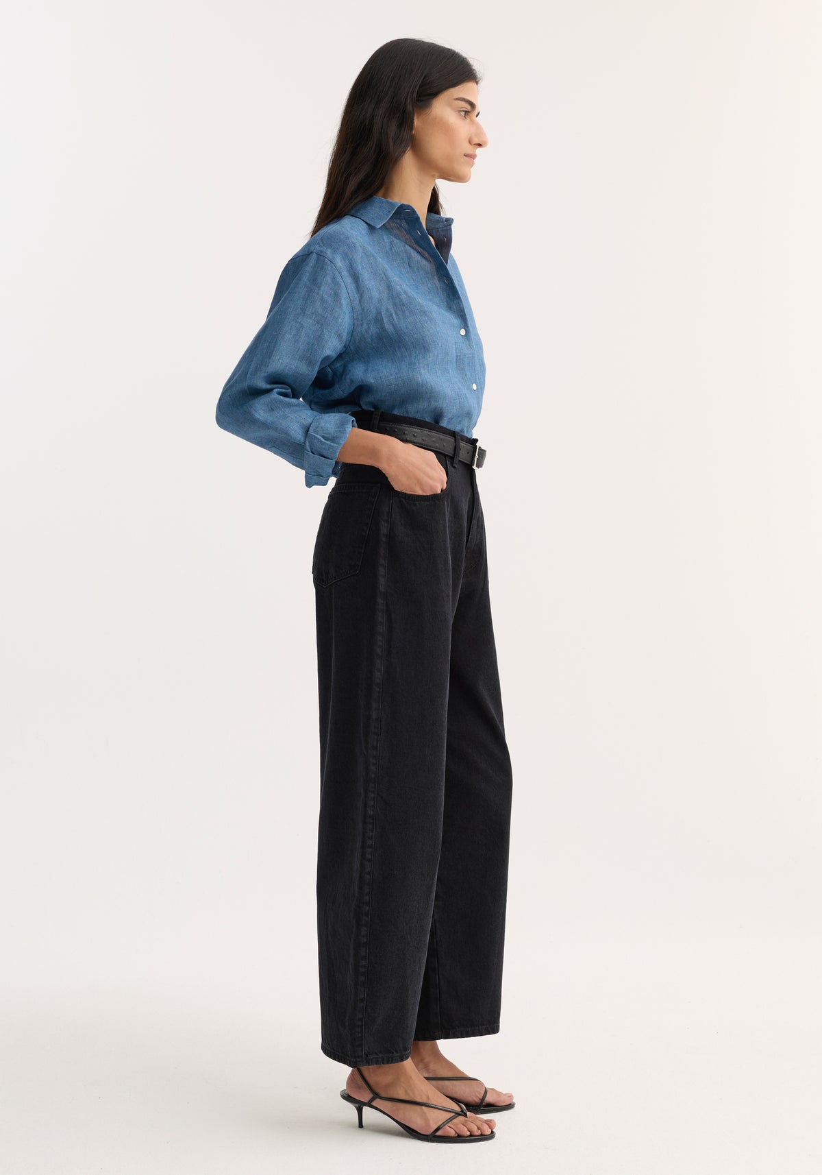 Woman wearing black Berber - Barrel leg denim | black pants paired with a blue button-up shirt and black open-toe heels, standing in profile against a white background.