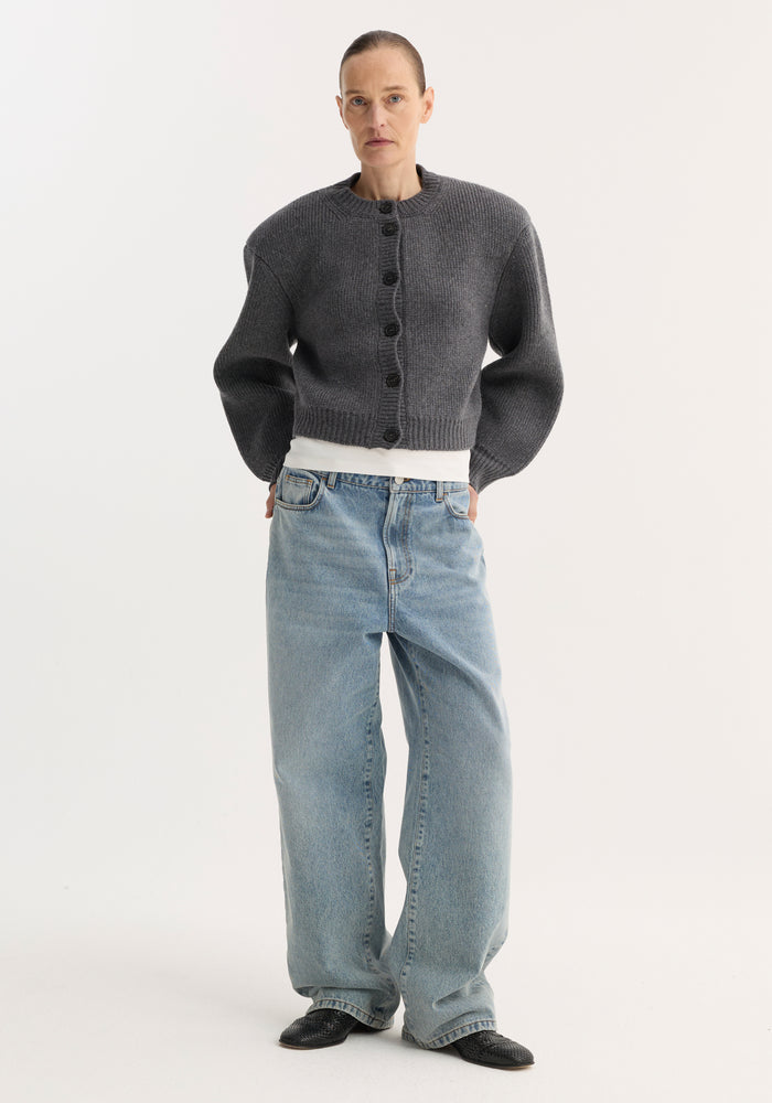 Person in charcoal cardigan and white T-shirt, wearing Signature baggy fit denim, classic blue, with wide-leg, low-waist fit, paired with black slip-on shoes.