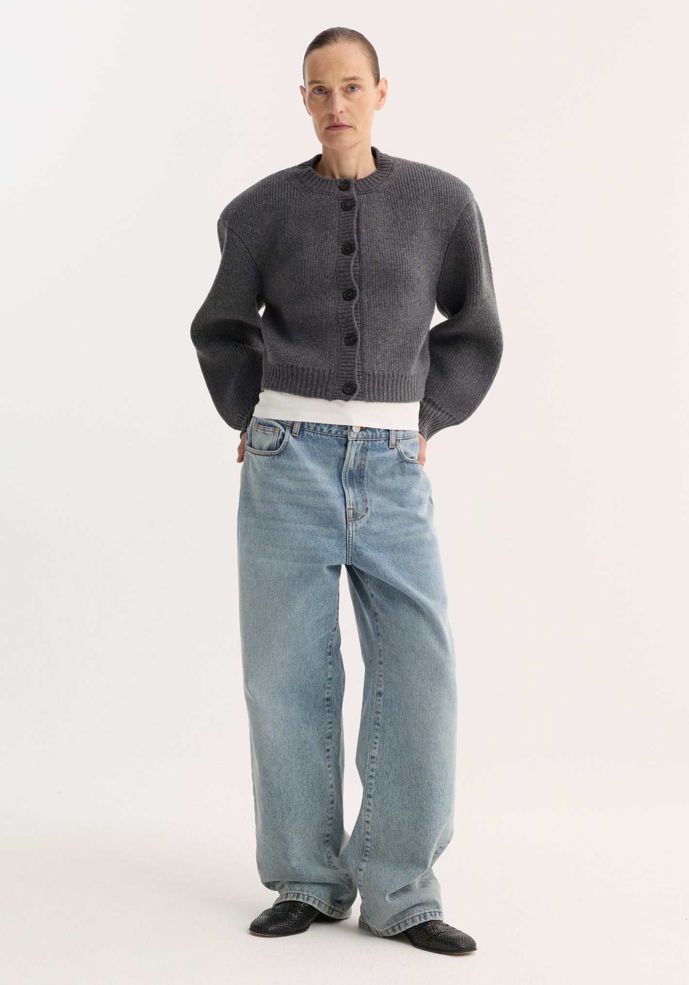 Person wearing Baggy fit denim | classic blue, low-waist, wide-leg jeans, paired with a gray cardigan, white T-shirt, and black mules.