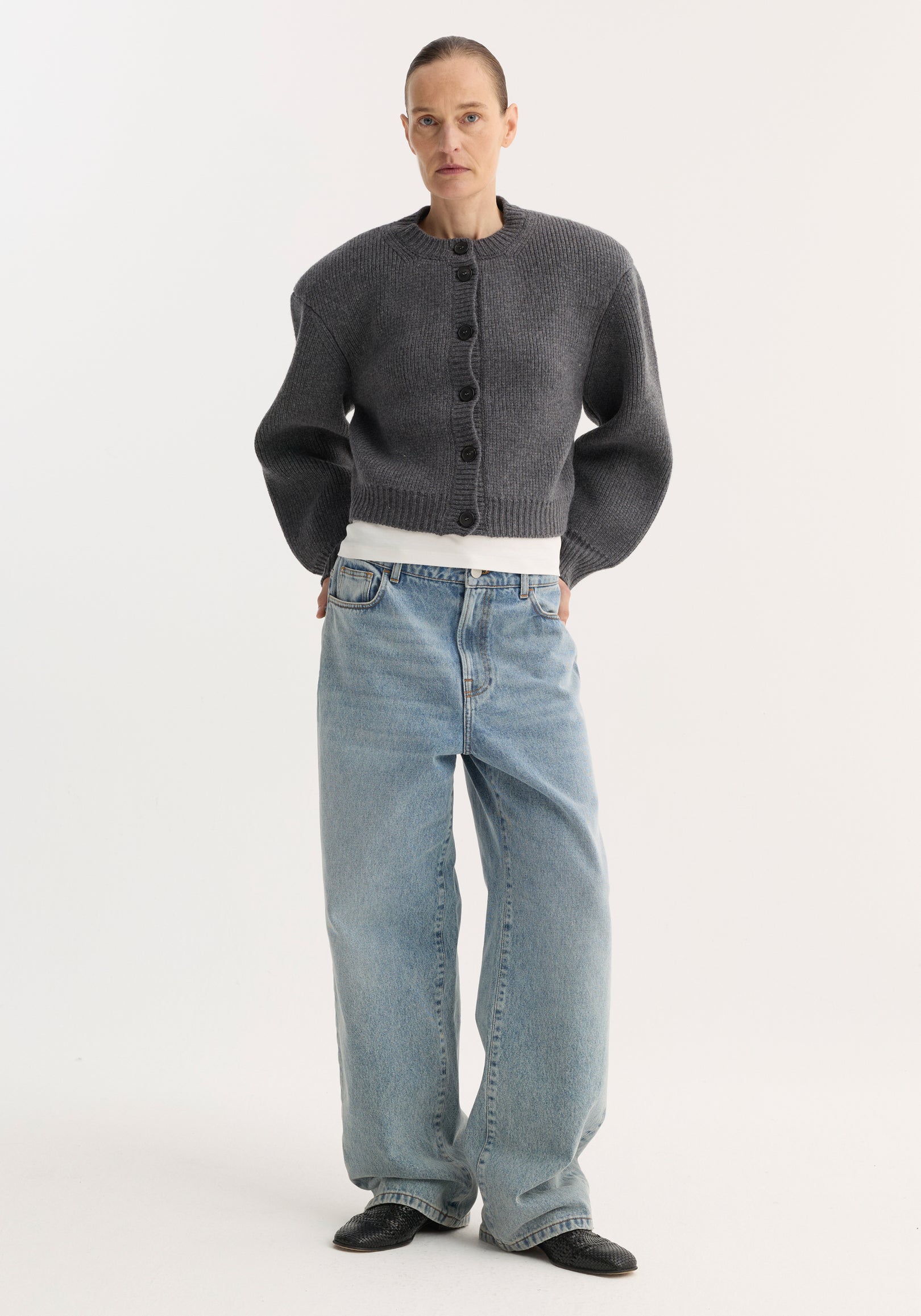 Person wearing Baggy fit denim | classic blue, low-waist, wide-leg jeans, paired with a gray cardigan, white T-shirt, and black mules.