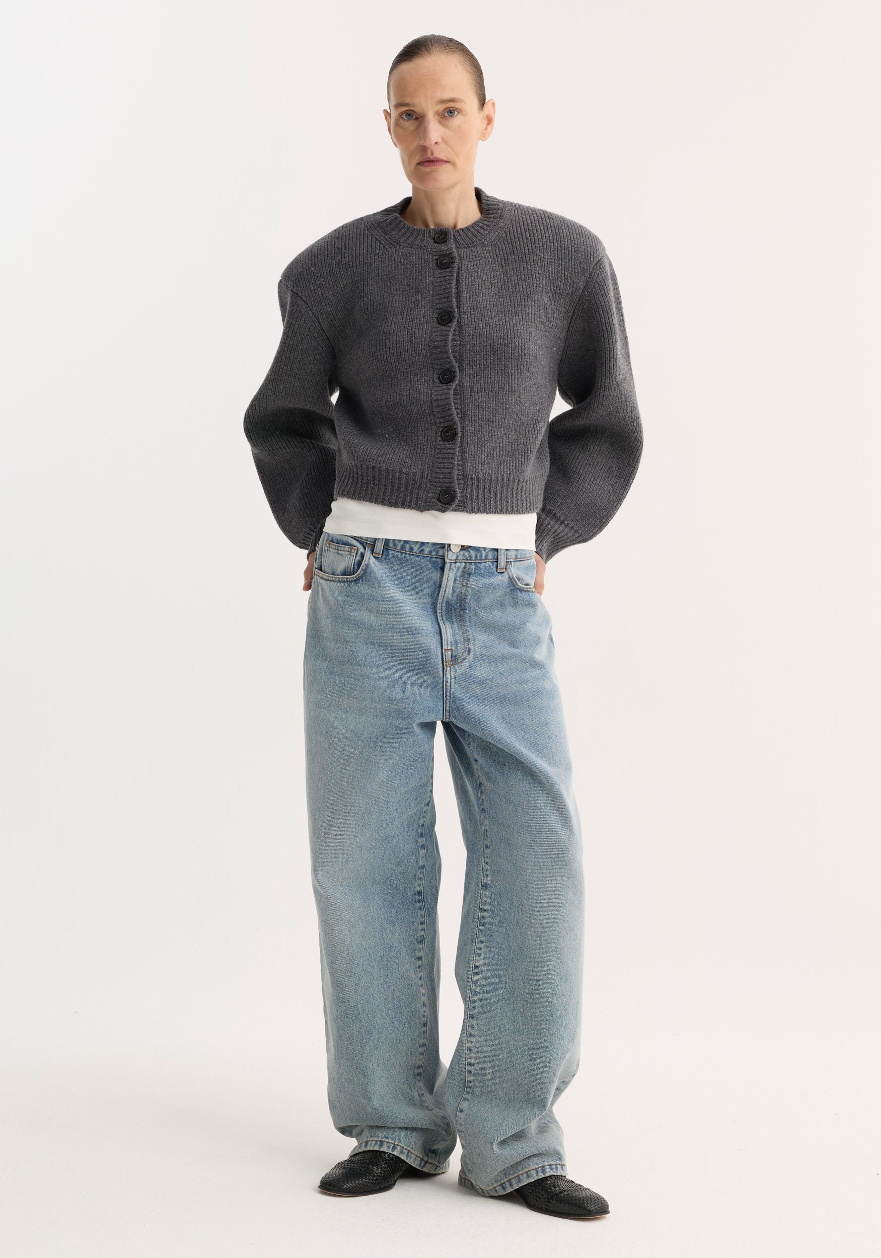 Person wearing Baggy fit denim | classic blue, low-waist, wide-leg jeans, paired with a gray cardigan, white T-shirt, and black mules.