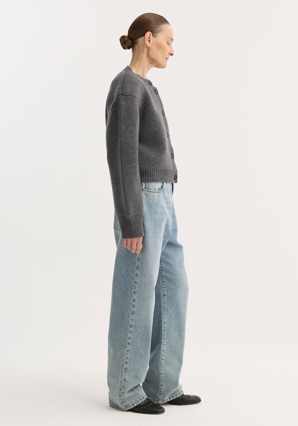 Side-profile of a person in baggy fit denim, classic blue, low-waist, wide leg, paired with a gray cardigan and black pointed-toe shoes.