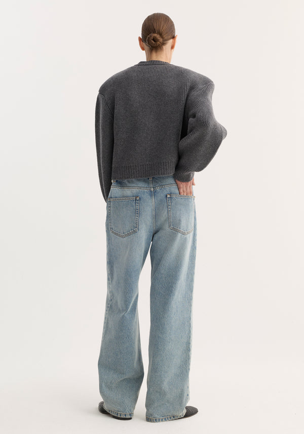 Person wearing Baggy fit denim | classic blue with patch pockets, ribbed charcoal sweater, and dark slip-on shoes, standing back to camera, hand in pocket.