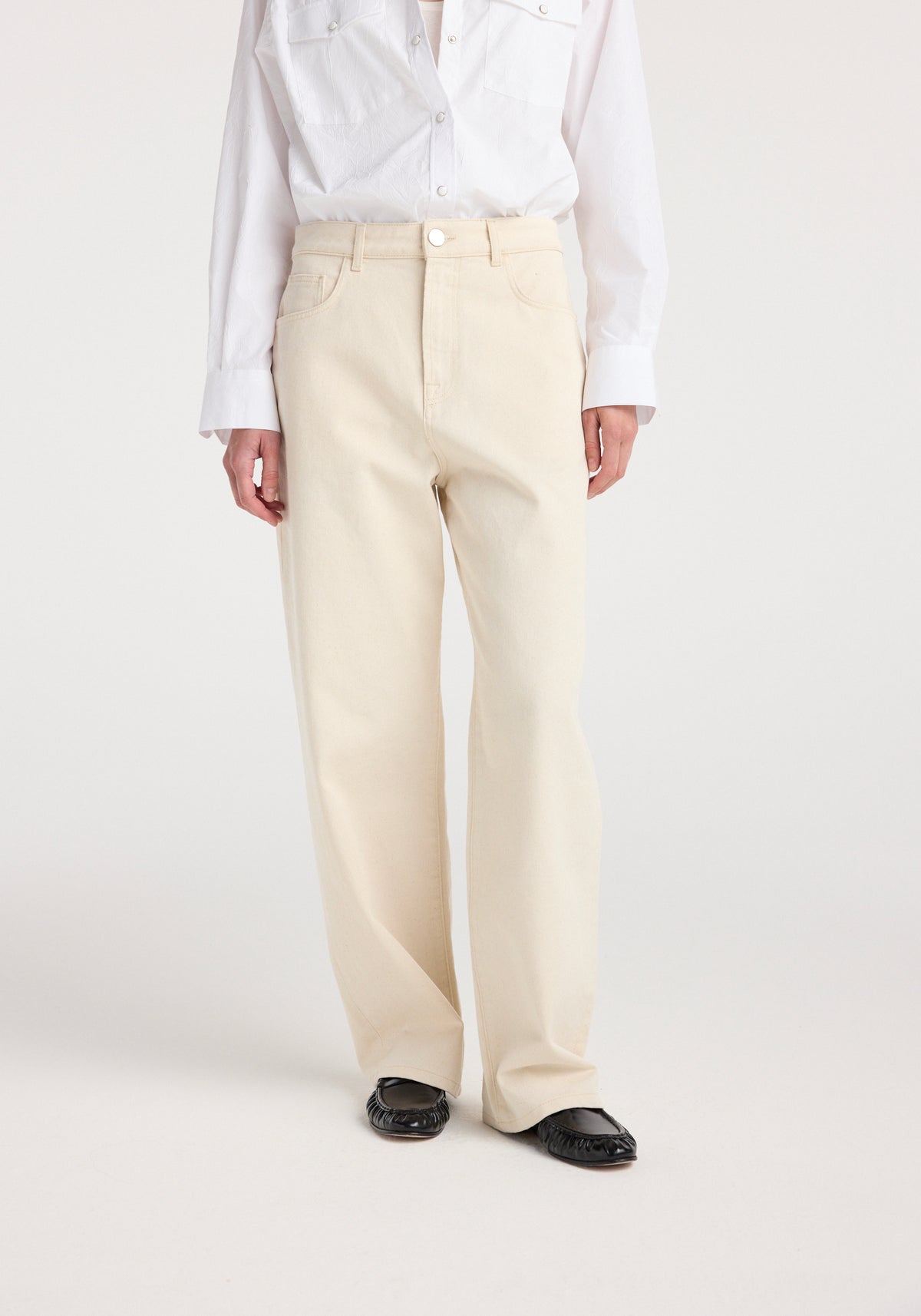 Front view of a person wearing ecru colored Bruun - Signature baggy denim paired with a white button-up shirt and black shoes against a plain background.