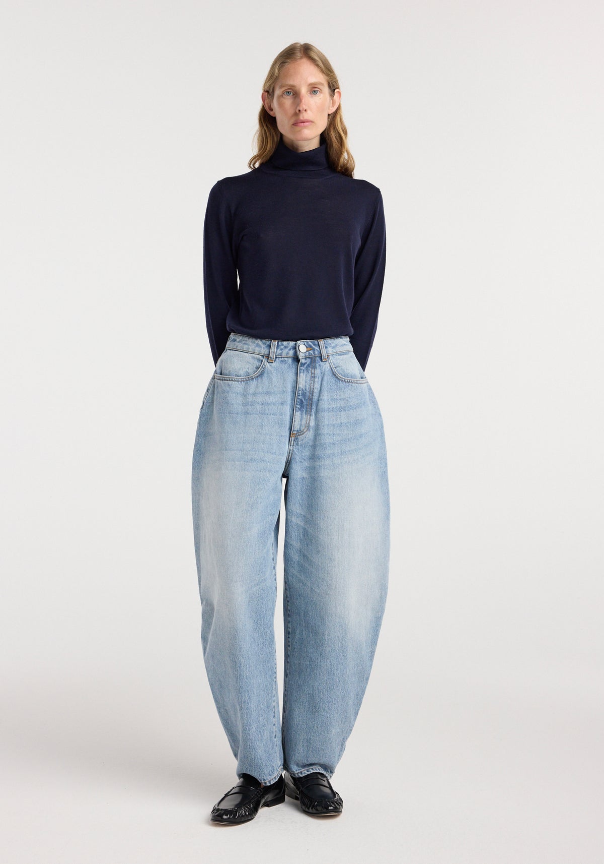 A front view of a model wearing Fransje - Sculptural denim trousers | classic blue, paired with a dark navy turtleneck and black shoes against a plain white background.